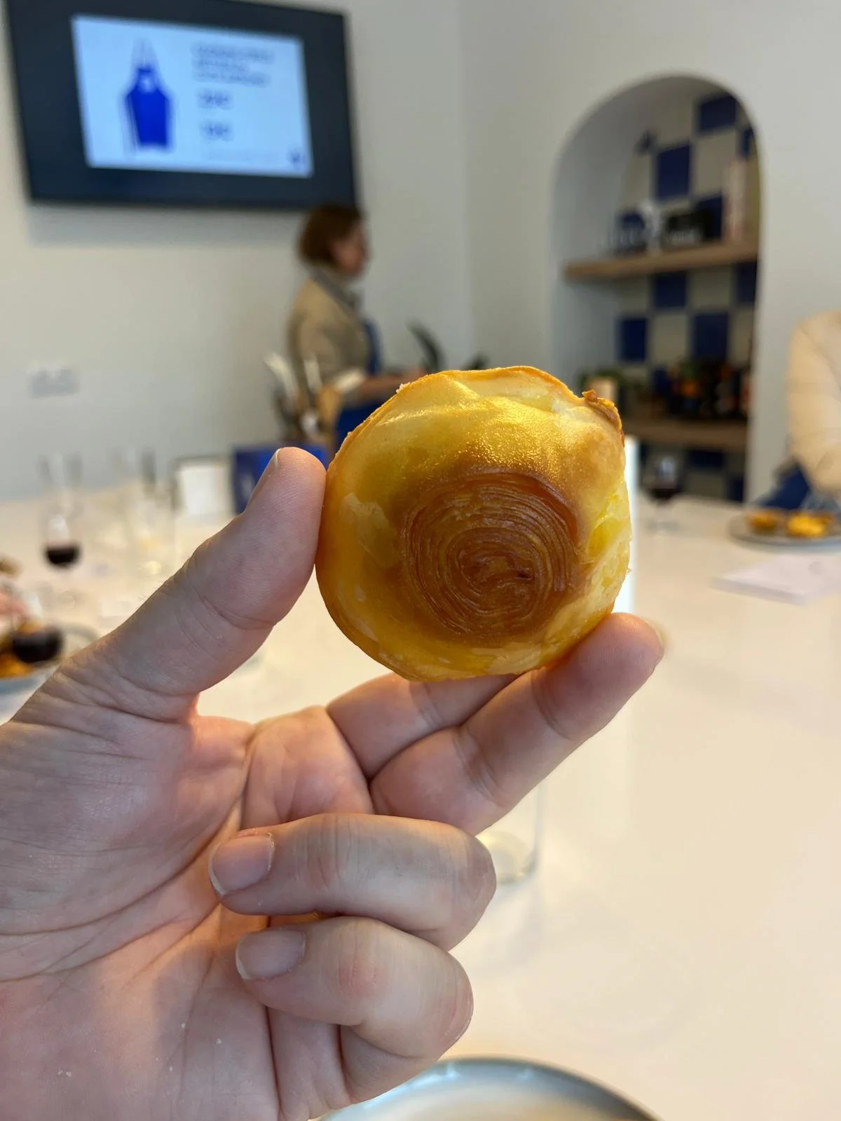 Close-up of a hand holding a pastry with a spiral pattern on top, in a room with a blurred background including a person, tables, glasses, and a television screen.
