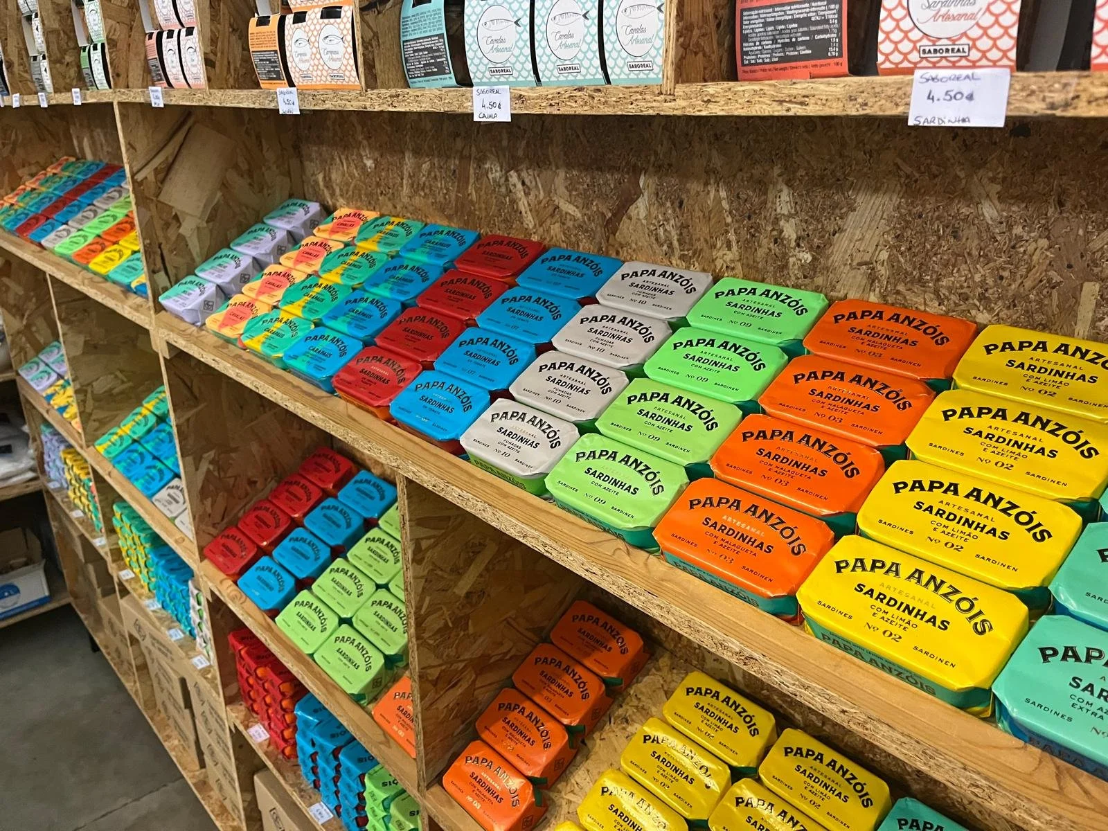 Shelves stocked with various brands and flavors of sardines in colorful packaging, including green, yellow, red, blue, and orange boxes.