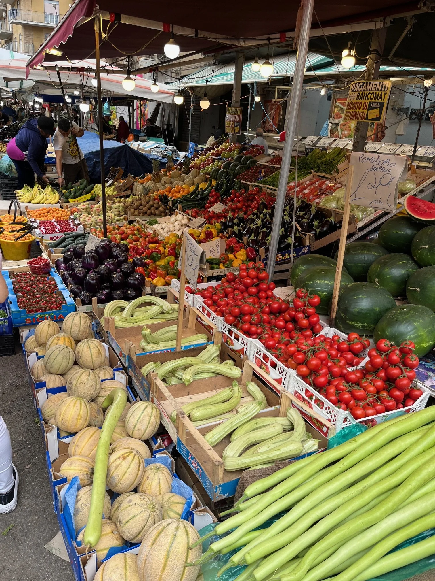 Fresh fruits and vegetables displayed in a  Portuguese market stall, including melons, squash, tomatoes, watermelon, eggplant, zucchini, and other produce, with people shopping and vendor signs in the background.