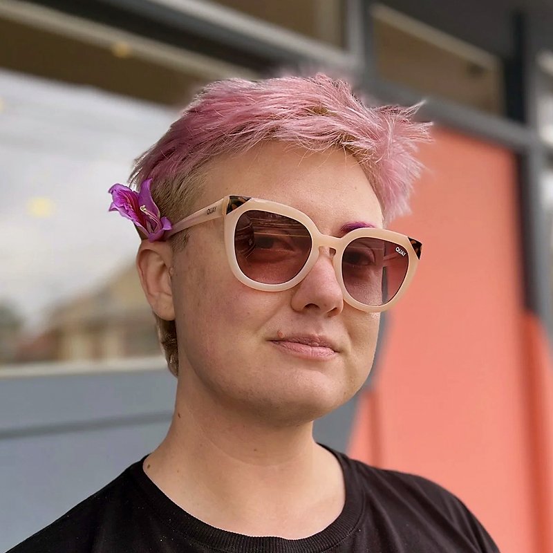 Dean, a white trans man with short pink hair, smiles softly, wearing peachy sunnies with a purple flower tucked behind his ear.