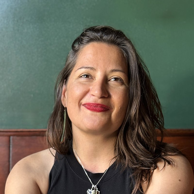 Lee, a white nonbinary woman with long brown hair, brown eyes, and wearing a popping red lipstick, smiles brightly in front of a green wall.