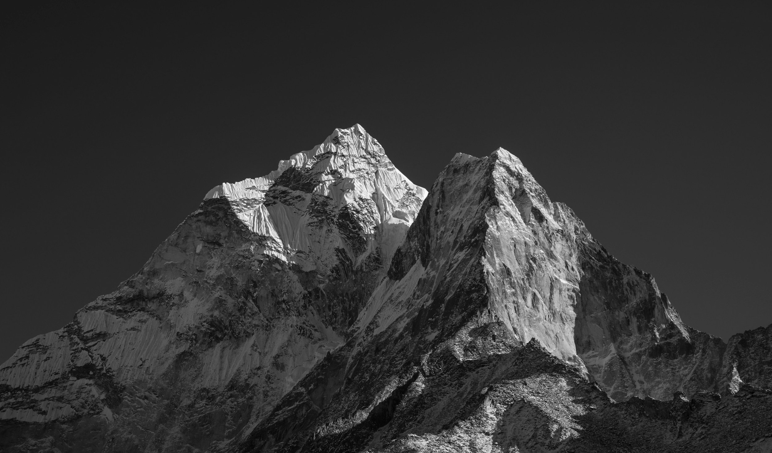 Black and white photograph of a tall mountain range with snow-covered peaks and rugged slopes against a clear sky.