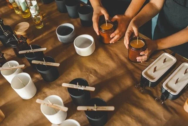 Hands holding jars of honey or jam over a wooden table with cups and bowls, possibly at a tasting or brewing event.