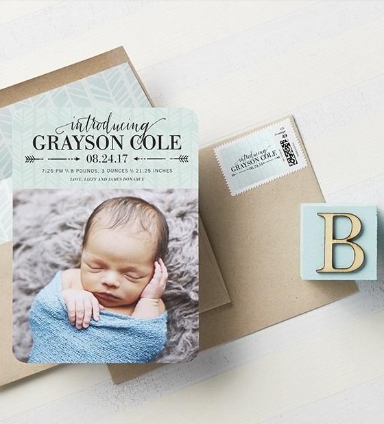 A birth announcement card with a baby sleeping, wrapped in a blue blanket, and the name Grayson Cole. The card includes details of birth date, time, weight, and length. It is placed on top of a brown envelope and a small box with a letter "B" on it.