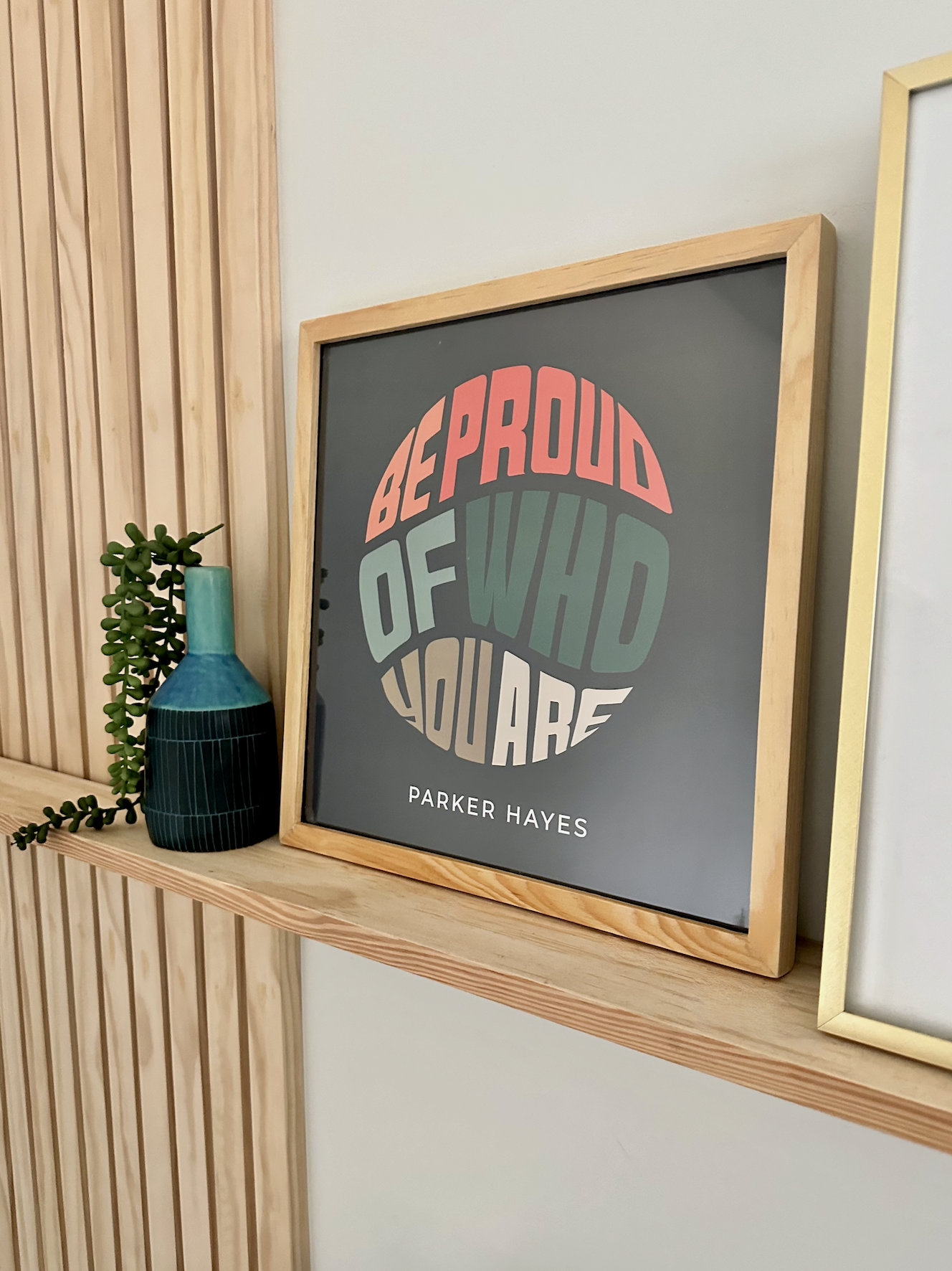 Decorative framed art with the phrase 'Be Proud of Who You Are' in colorful block letters on a dark background, placed on a wooden shelf next to a small green plant and a blue and green vase.