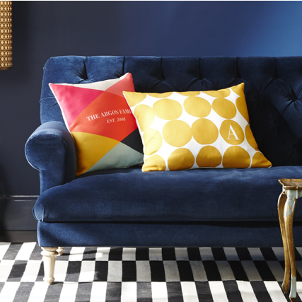 Blue sofa with two decorative pillows, one with a colorful geometric pattern and the other with yellow circles and white initials, a black and white checkered rug, and a dark wall with a window.