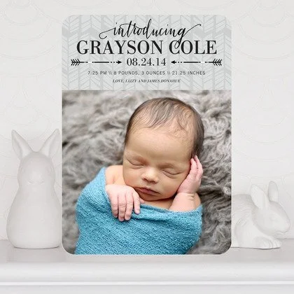 Baby sleeping wrapped in a blue blanket with hands near face, baby shower announcement card above, white bunny figurines on each side.