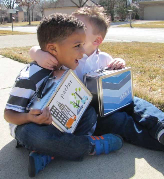 Two young boys sitting on the sidewalk, hugging and smiling. They are holding lunchboxes, one with a school bus and tree design labeled 'Parker,' and the other with a blue geometric pattern labeled 'Jack F.' They are outside on a sunny day.