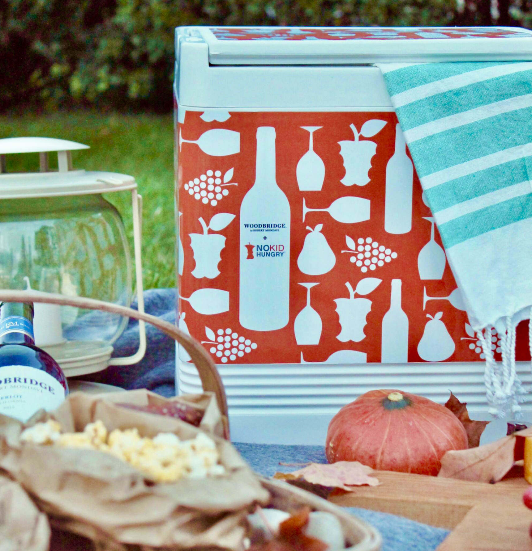 A red and white cooler with fruit and bottle patterns, partially covered by a blue and white striped cloth, next to a pumpkin and wood tray with food, set outdoors on grass.