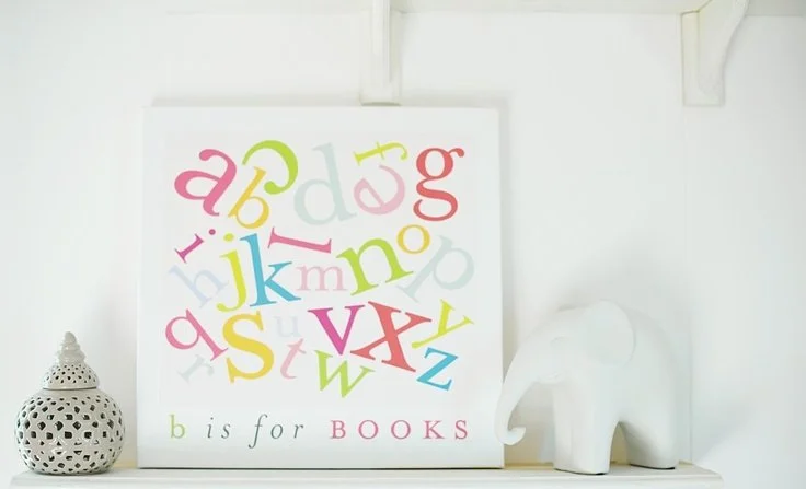 Decorative alphabet print with various colorful letters and the phrase "b is for BOOKS", placed on a white shelf alongside a white ceramic lantern and a white elephant figurine.