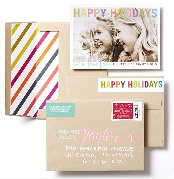 A beige envelope with a colorful striped card to the left. The envelope has a green address label, a pink handwritten name, and a red holiday stamp. A black-and-white photograph of three smiling women and children with a 'Happy Holidays' banner and a