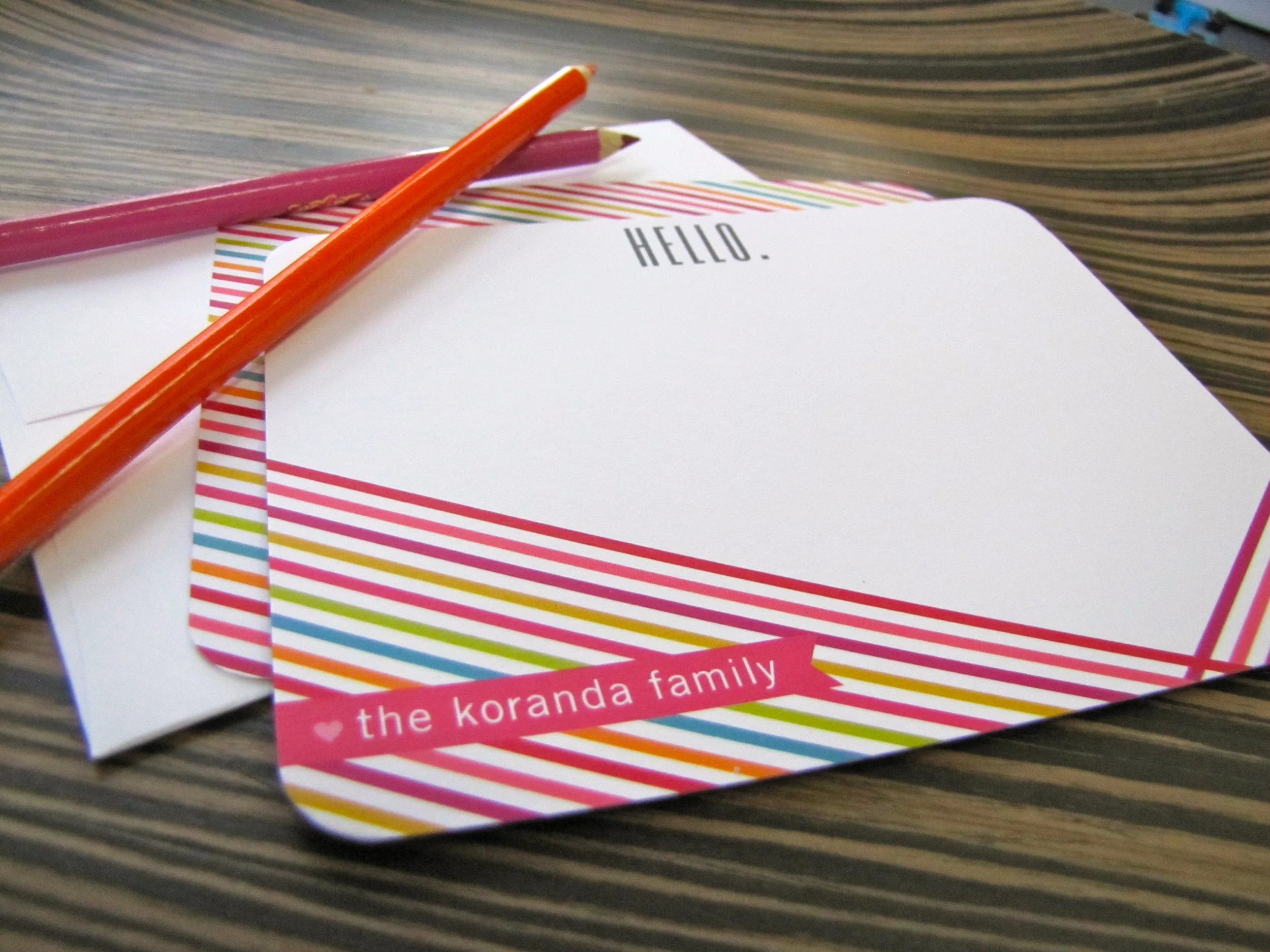 Colorful striped cards with the text 'HELLO.' and a small banner with 'the koranda family,' along with a pink pencil and an orange pencil on a striped table.