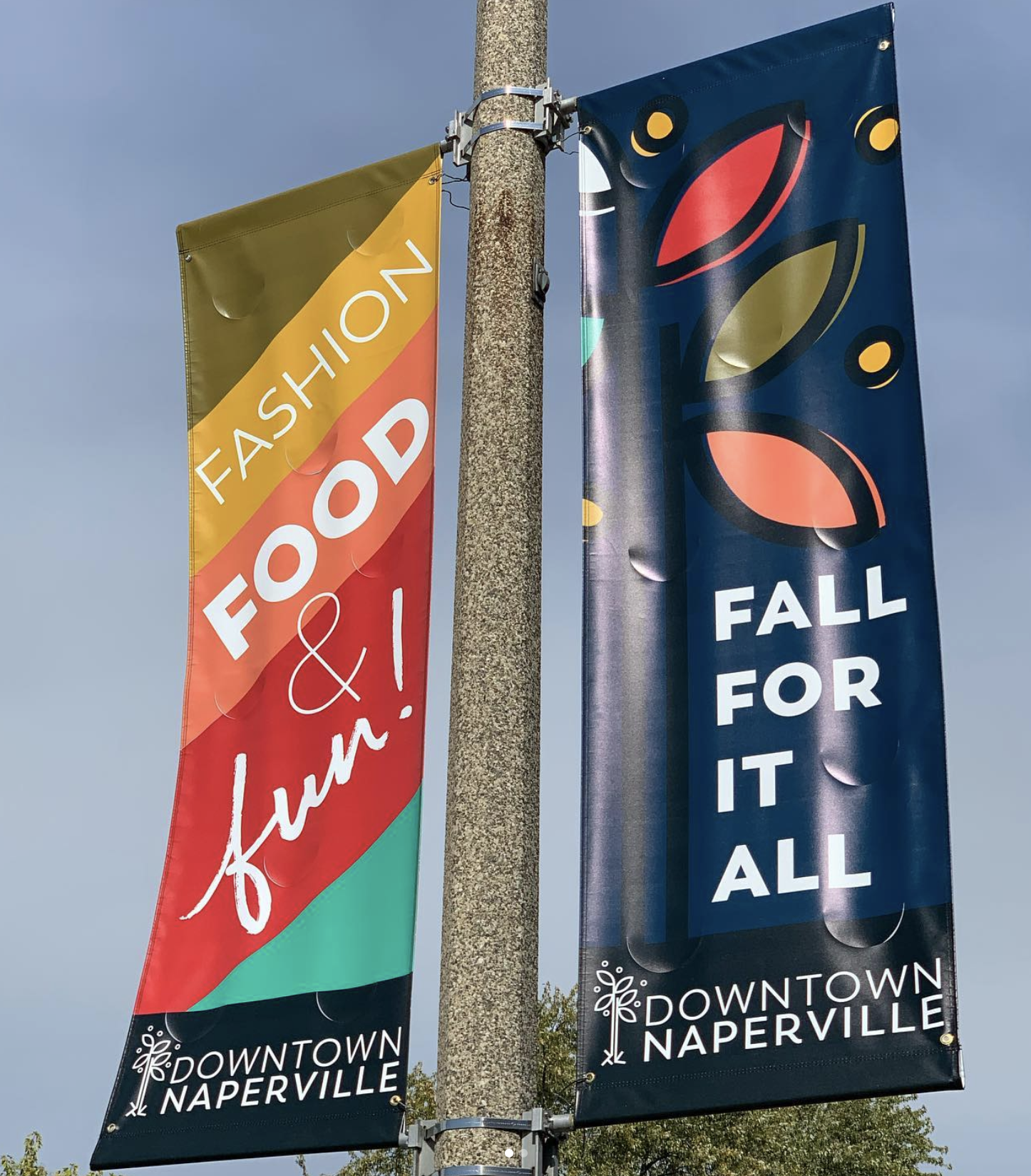 Two colorful vertical banners hanging from a pole in downtown Naperville. The left banner promotes a fashion food and fun event, and the right banner encourages falling for it all.