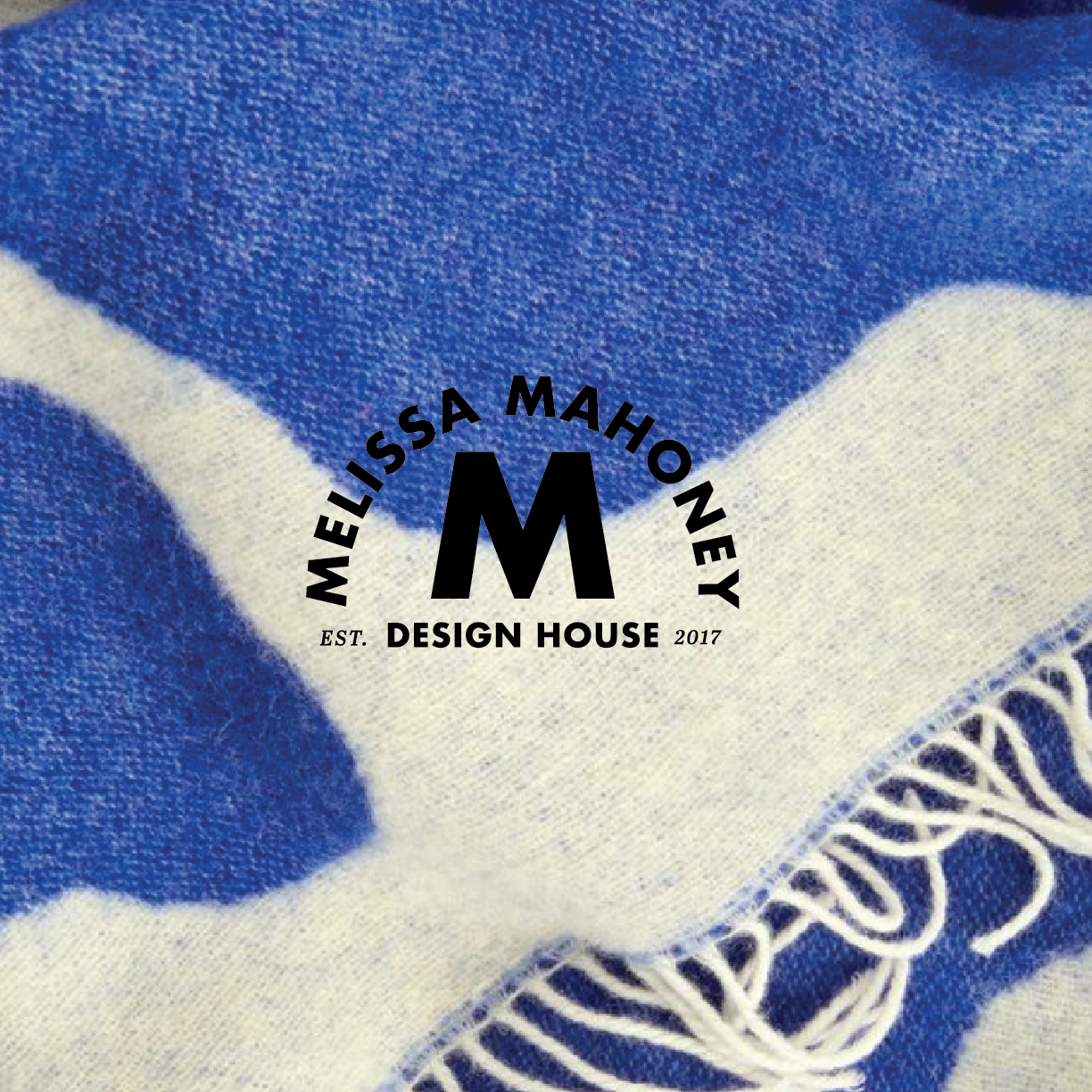 Close-up of fabric with the designer logo, 'Melissa Mahoney Design House' and a large 'M' in the center, on a blue and white patterned cloth.