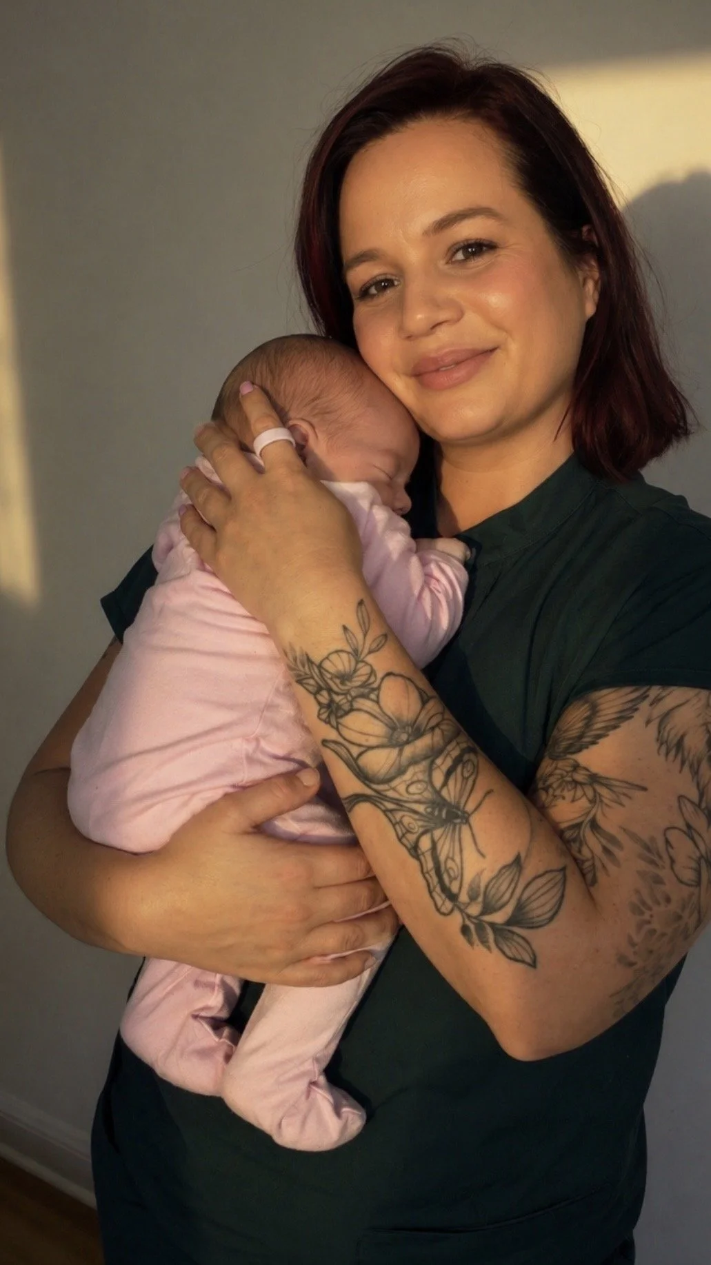 A woman with short brown hair and tattoos on her arms holds a sleeping baby in a pink outfit, gently resting her face on the baby's head.