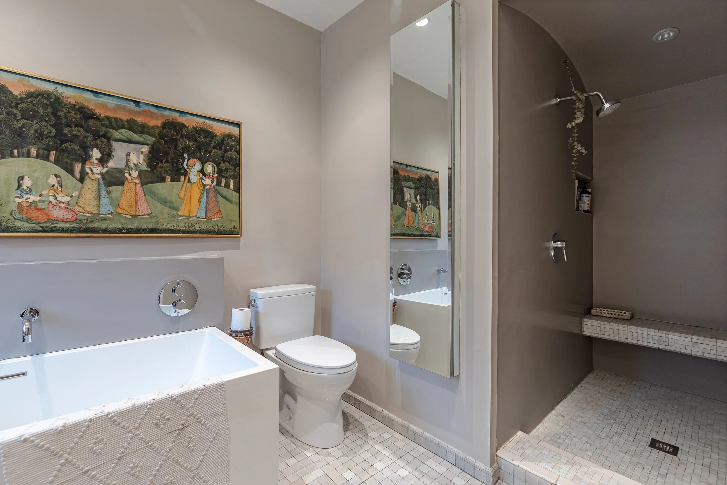 A modern bathroom with a white bathtub, toilet, large mirror, wall art depicting a scene with women in traditional clothing, and a walk-in shower with a bench and handheld shower head.