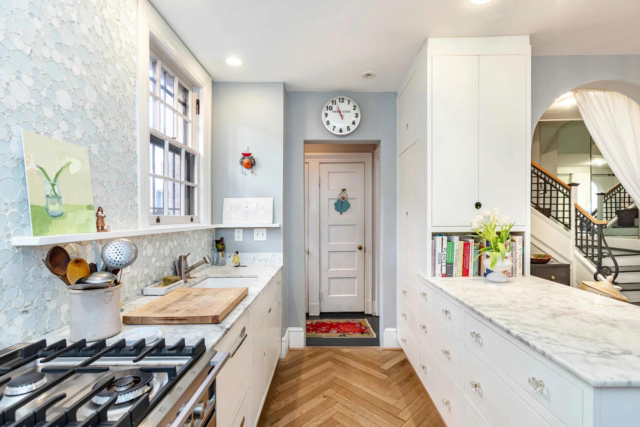 Bright kitchen with white cabinets, marble countertops, a gas stove, a window with a ledge holding artwork, a clock showing 5:14, decorative items, and a view into a staircase area.
