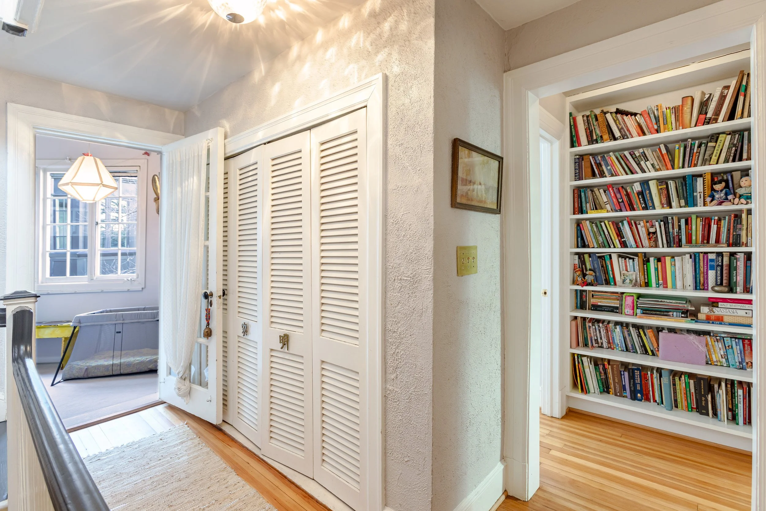 A hallway with white louvered closet doors, a framed picture on the wall, and a door leading to a room with a bookshelf filled with books.