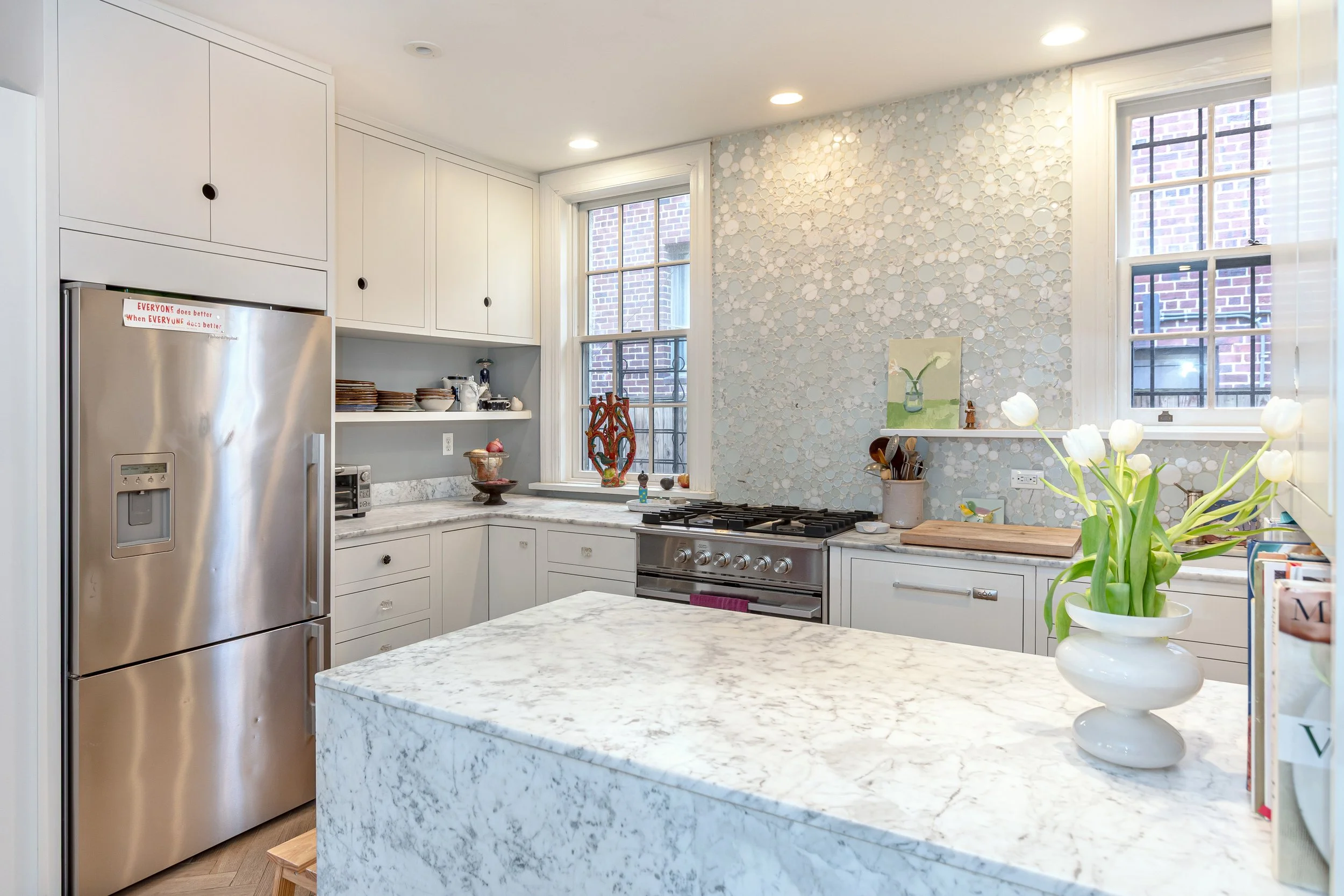 Modern kitchen with white cabinetry, marble island, stainless steel refrigerator, windows with brick buildings outside, decorative wall, and flowers in a white vase.