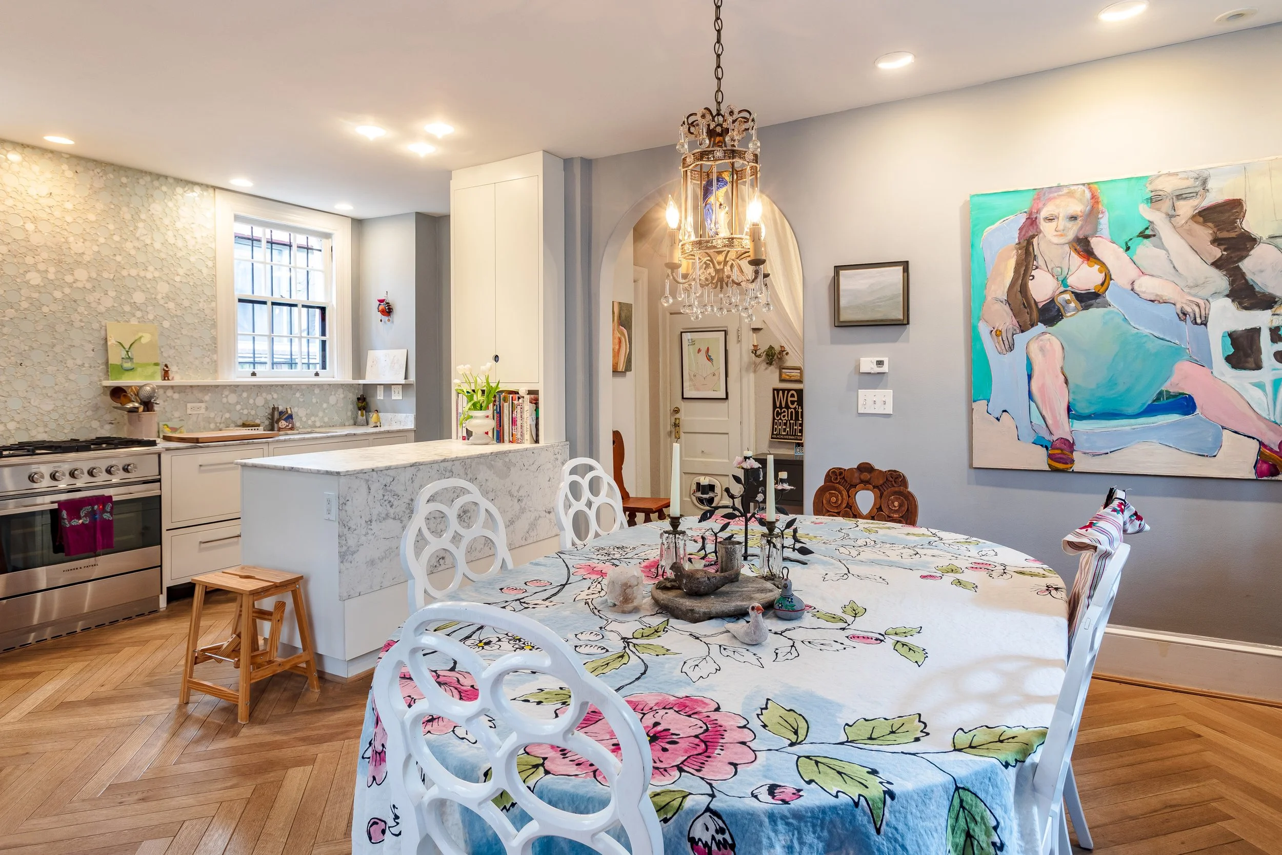 A dining area adjacent to a kitchen with a floral tablecloth, white chairs, and a chandelier. The kitchen features white cabinets, a marble island, and art decorations on the walls.