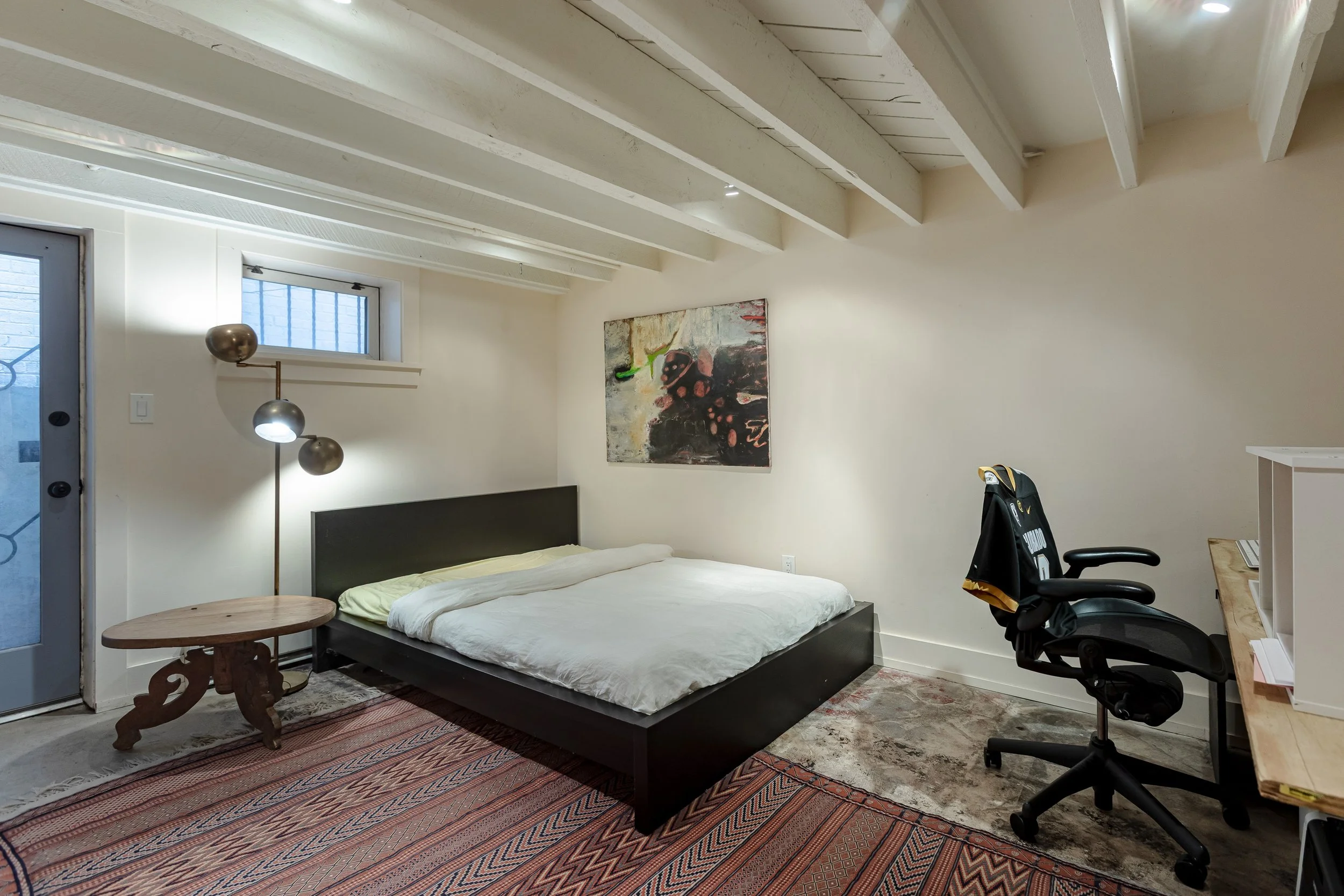 A bedroom with a black platform bed, a wooden side table, a modern floor lamp with three spherical shades, an abstract painting on the wall, a black office chair, a wooden desk with a computer, a small window, and a door with a decorative security sc