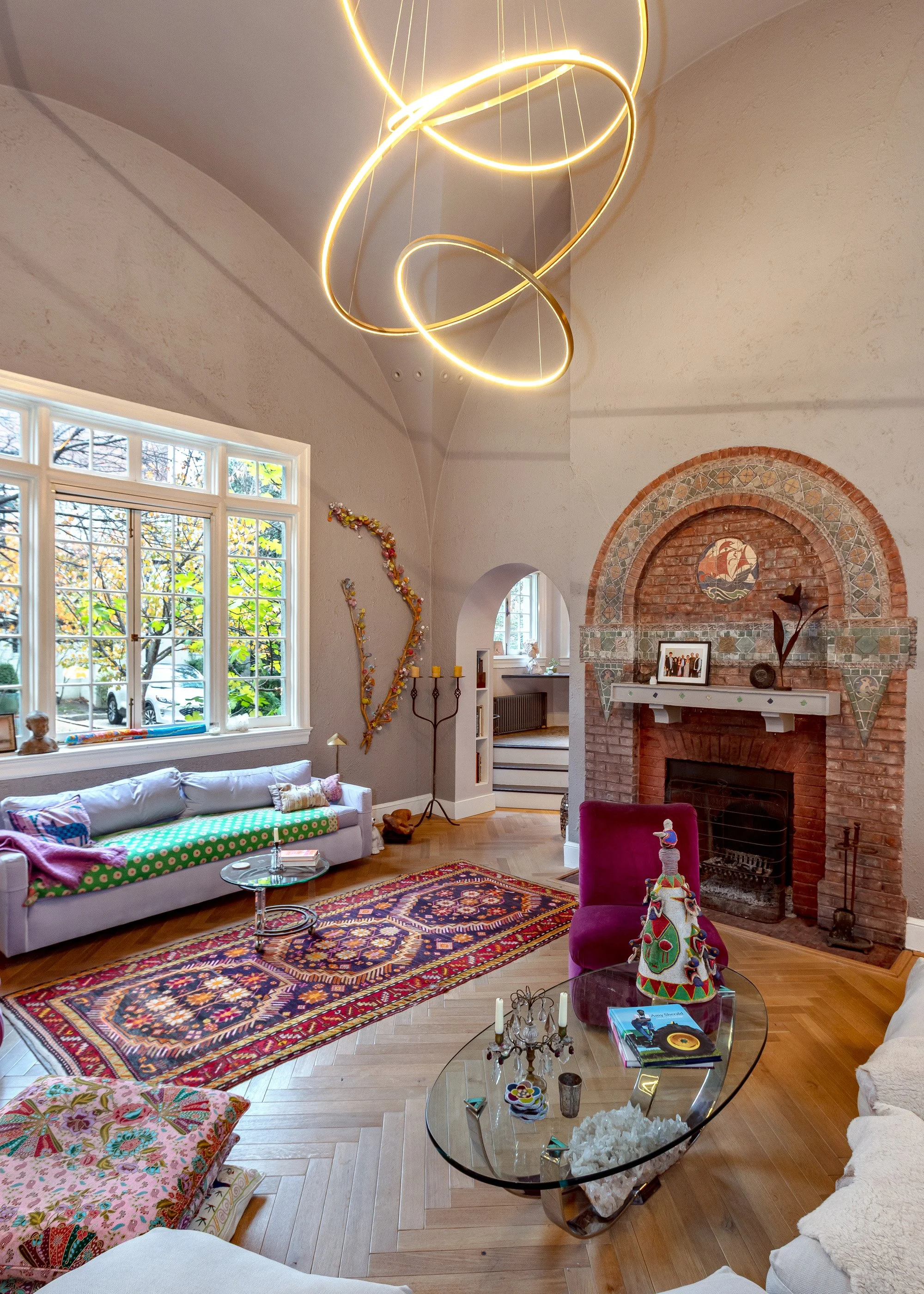 Living room with a high vaulted ceiling, modern gold chandelier, large window, white sofa with colorful pillows, patterned rug, brick fireplace, and eclectic decorations.