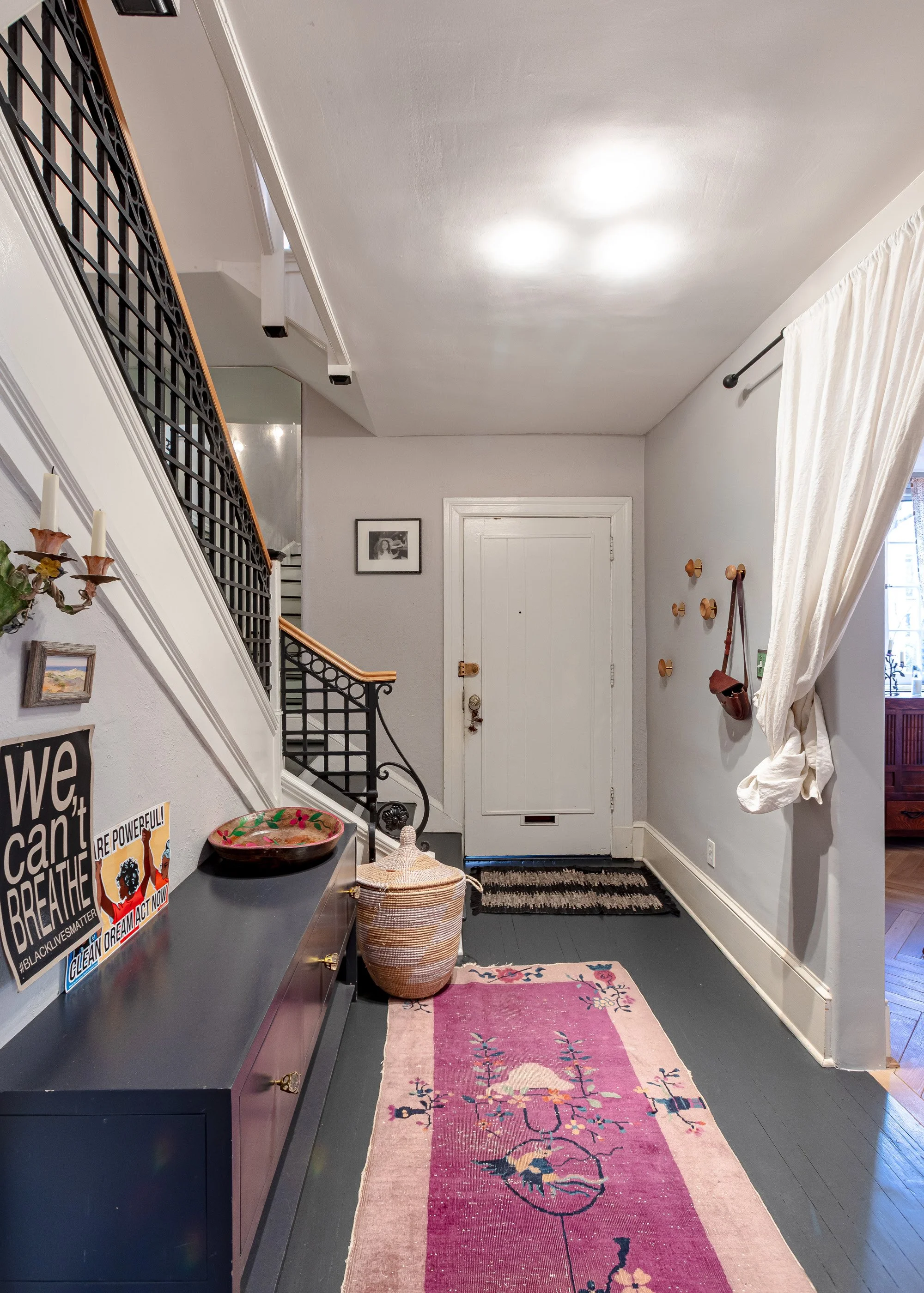 Interior entryway with front door, colorful pink and blue rug, side table with baskets, wall hooks, window with curtain, staircase railing.