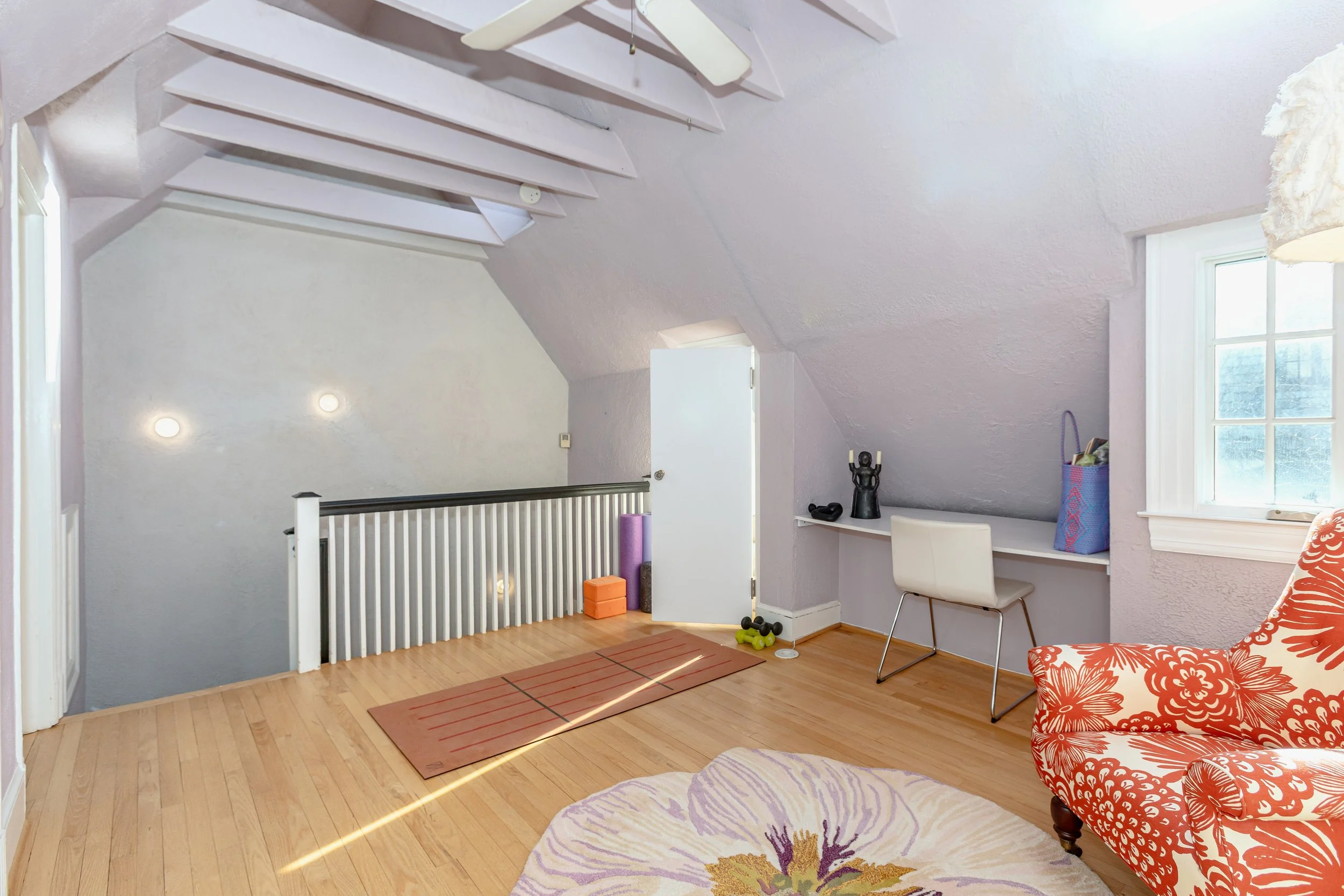 A cozy attic room with purple walls, hardwood floors, and a sloped ceiling with exposed beams painted white. There is a small open door, a white desk with a white chair, and orange and purple yoga mats and weights on the floor. A black sculpture and 