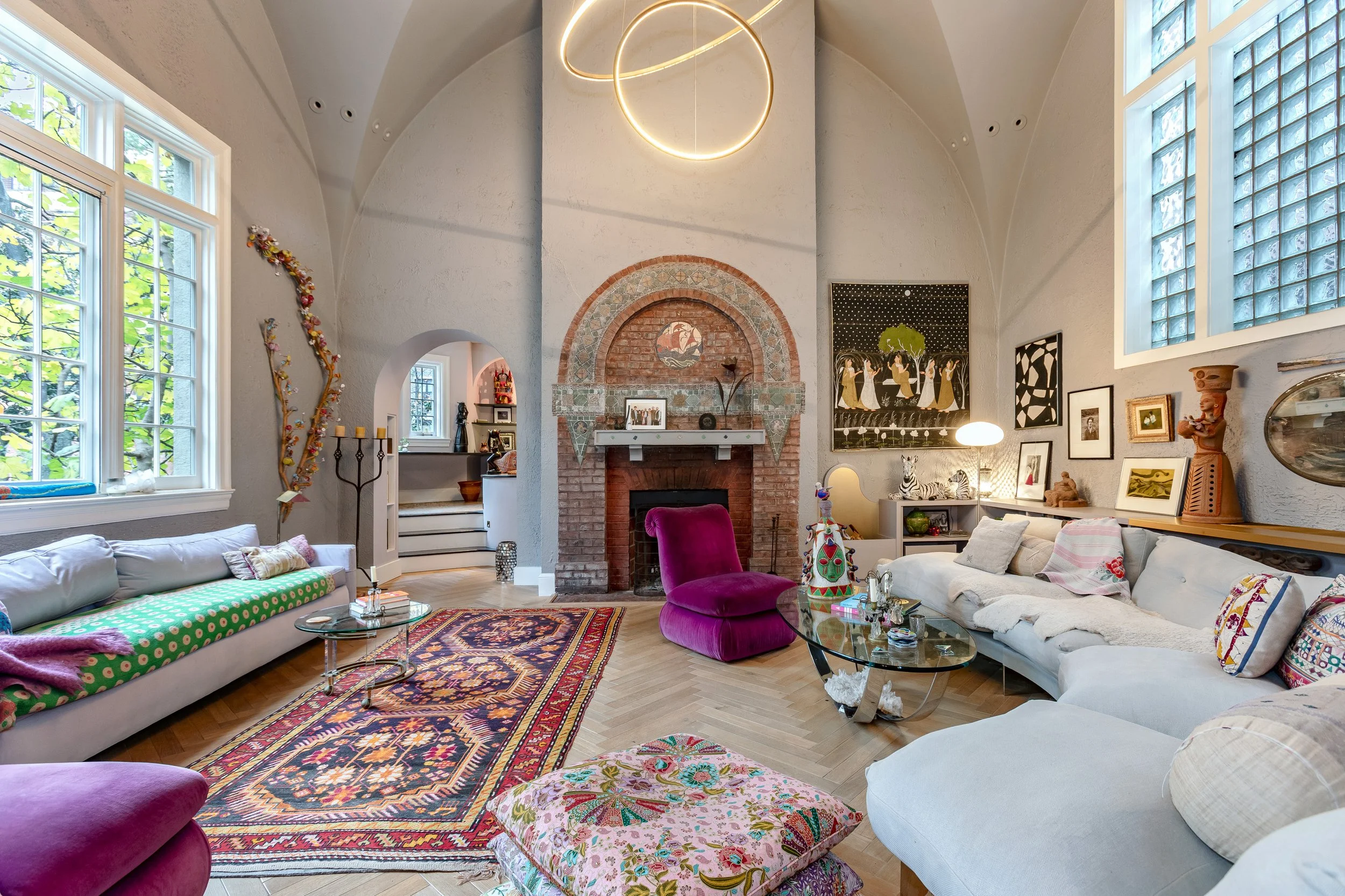 A cozy living room with eclectic decor, including a white sofa, colorful pillows, a glass coffee table, a pink velvet chair, and a patterned rug. There is a fireplace with an arched brick design, large windows with greenery outside, and modern circul
