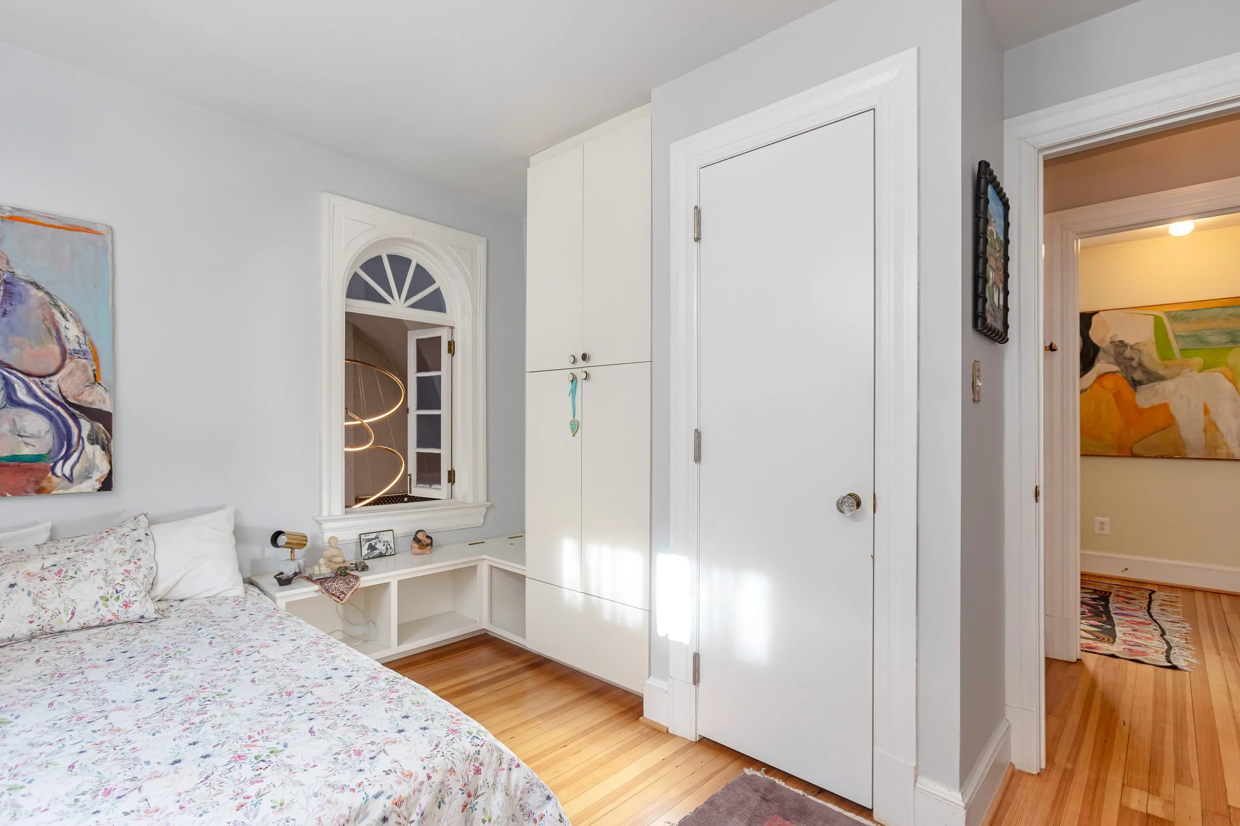 A bedroom corner with a bed, floral bedding, a white built-in storage unit, a small window with a curved top, and an open door leading to a hallway with a colorful abstract painting on the wall.