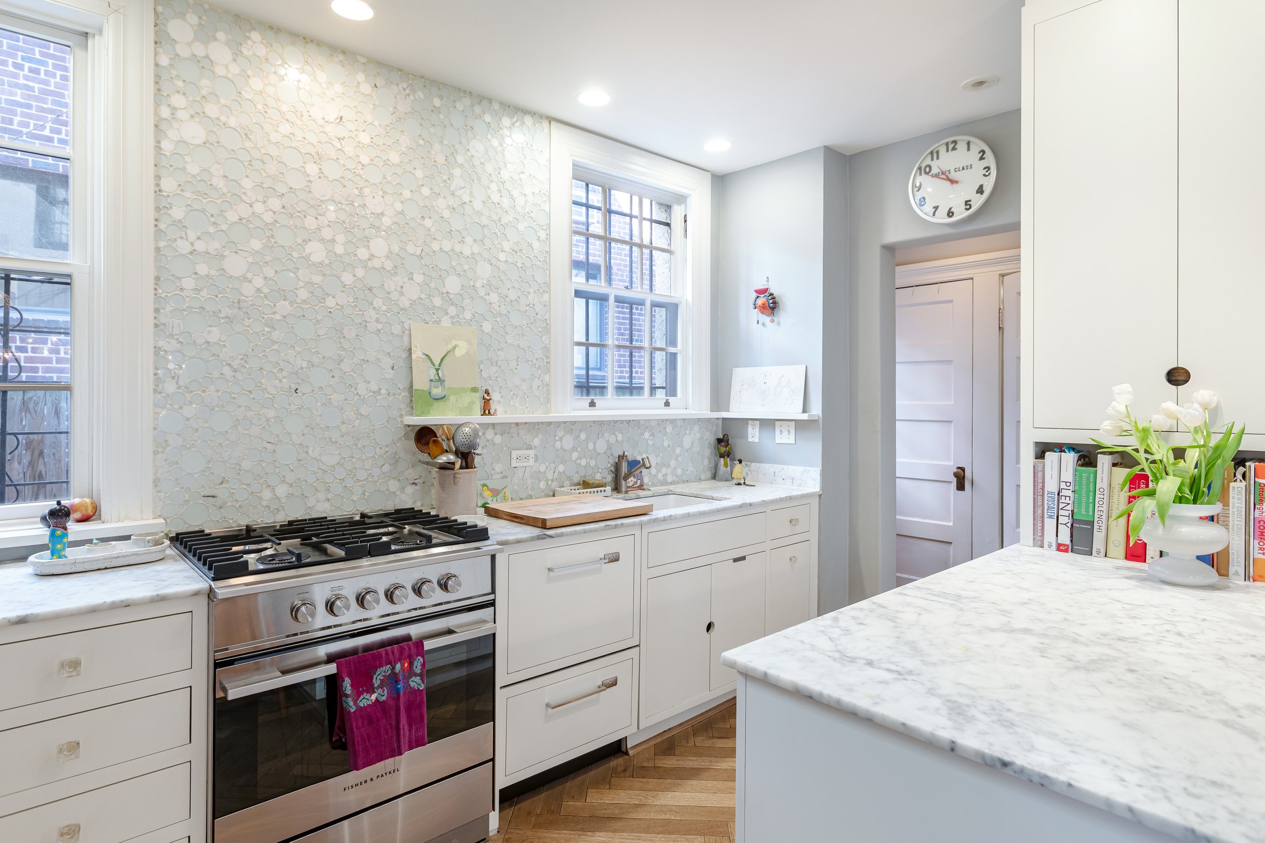 Bright kitchen with white cabinets, a marble countertop, and a mosaic tile backsplash. Two windows allow natural light, with various kitchen items and a small bookshelf with cookbooks on the counter. A clock on the wall shows 2:48.