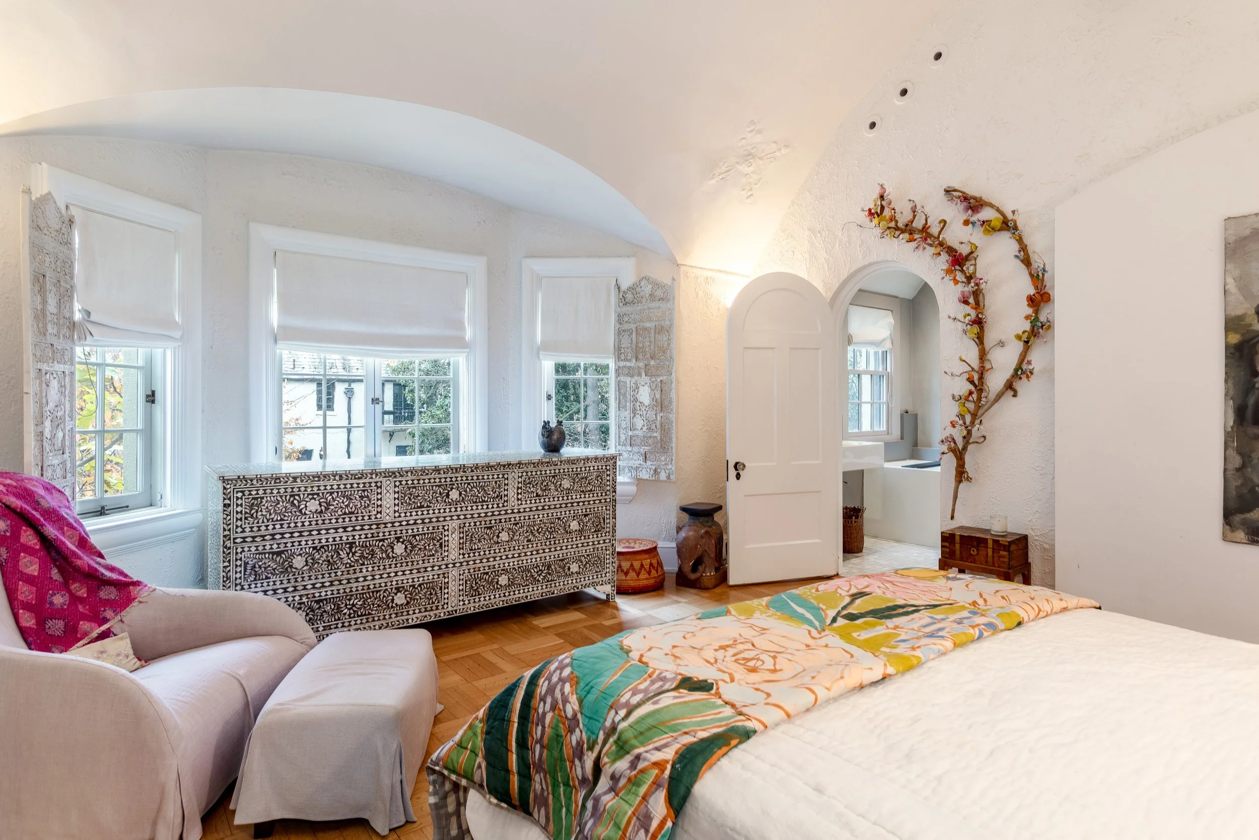 Bedroom with three windows, white walls, a bed with a colorful floral quilt, a white armchair with a pink throw, a luggage chest set, a small windowed bathroom with a bathtub, and decorative art including a branch sculpture.