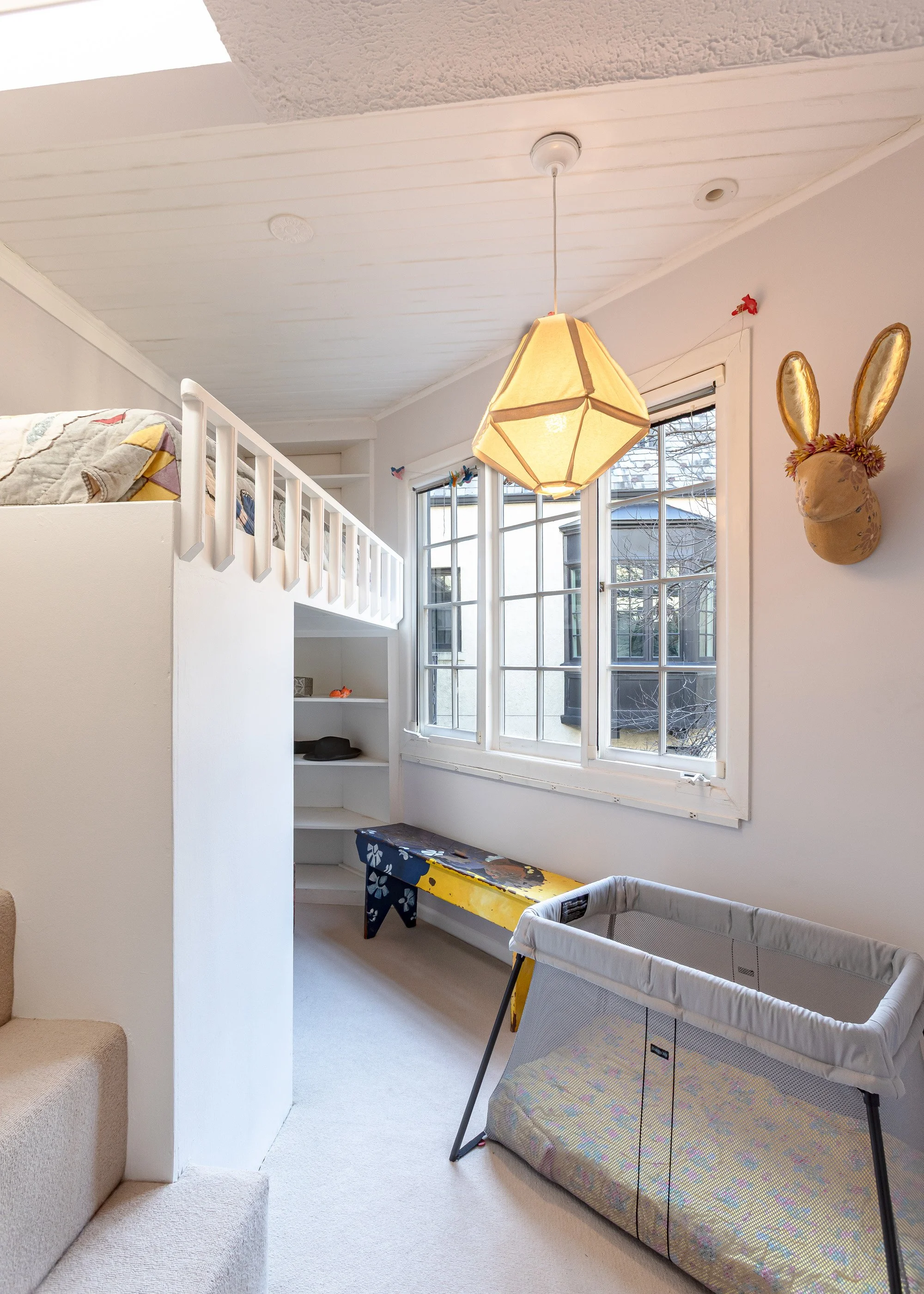 Children's bedroom with white loft bed, bookshelf, bench under window, hanging bunny wall decor, and changing table.