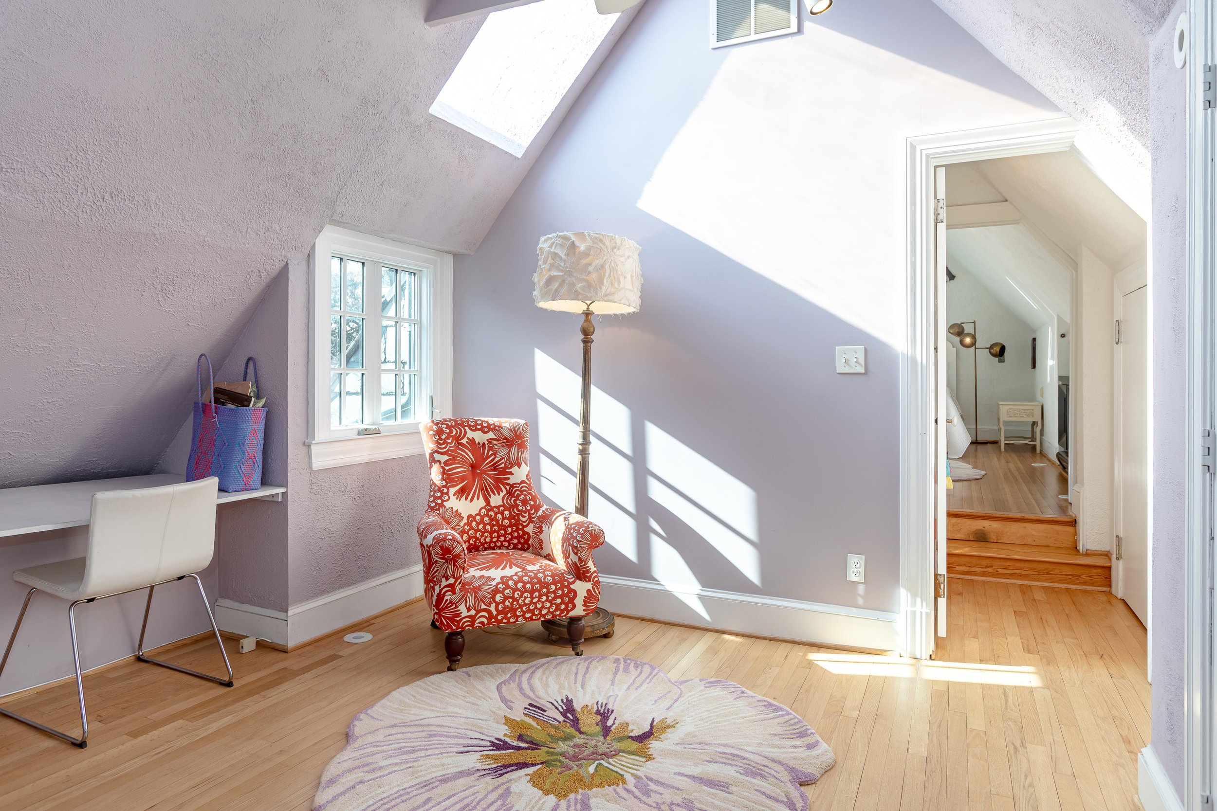 Bright attic room with sloped ceiling, skylight window, small rectangular window, white walls, hardwood floor, floral area rug, red patterned armchair, white desk with purple bag, standing lamp, and open doorway leading to another room with wooden st