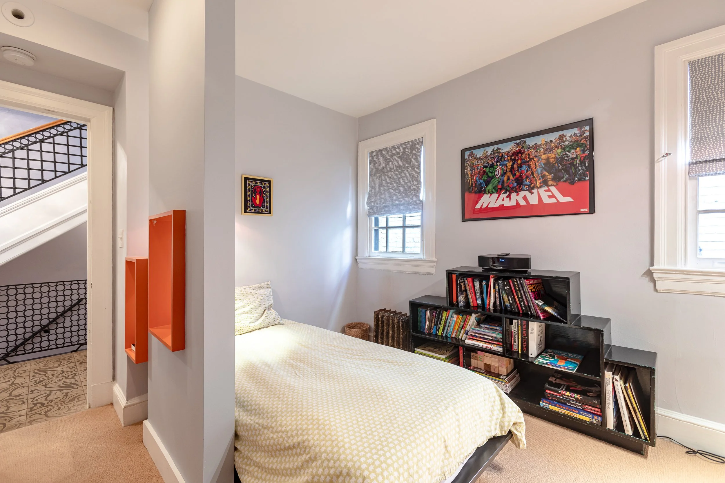 Bedroom with a bed, a black bookshelf filled with books, Marvel poster on the wall, two windows with blinds, and decorative shelving on the wall.