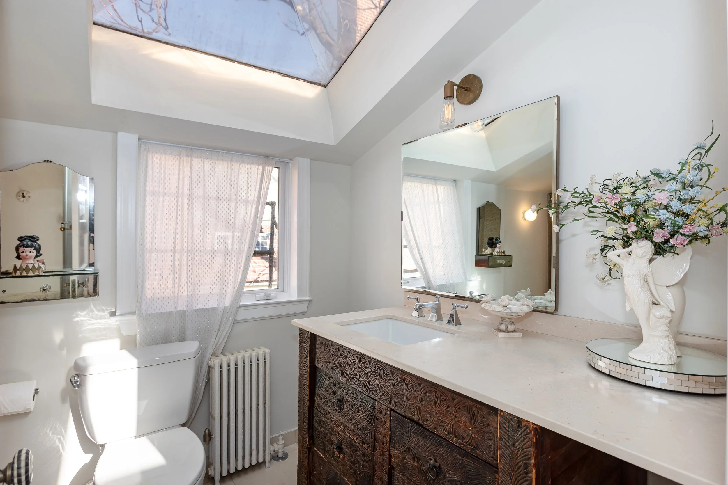 Bathroom with white walls, a small window with sheer curtains, a large mirror above a beige countertop with a sink, decorative items including a vase with flowers, a small shelf with figurines, and artwork on the ceiling.