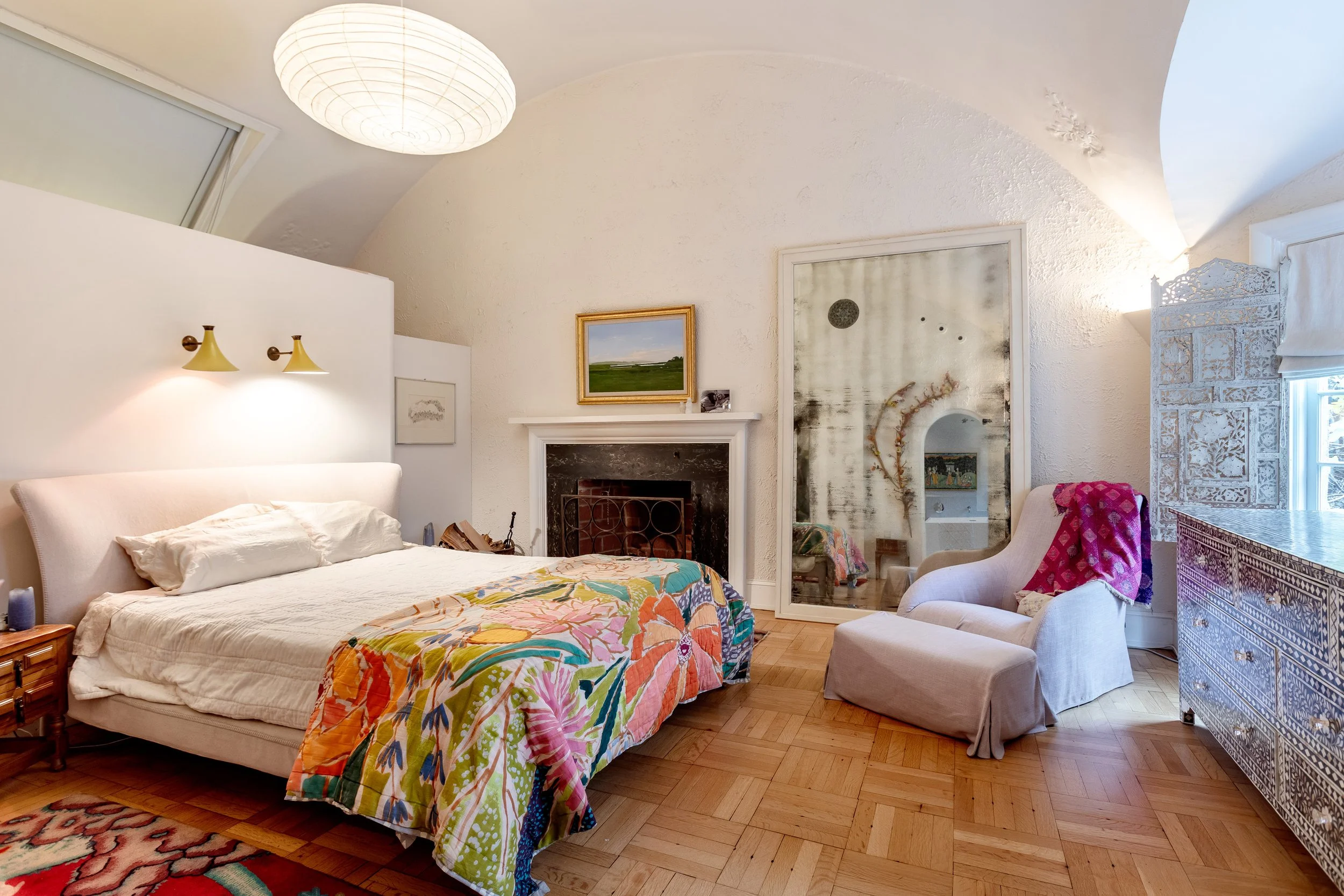 A cozy bedroom with a white bed, colorful quilt, wooden nightstand, fireplace, armchair with a pink blanket, mirrored wardrobe, window, and wooden floor.