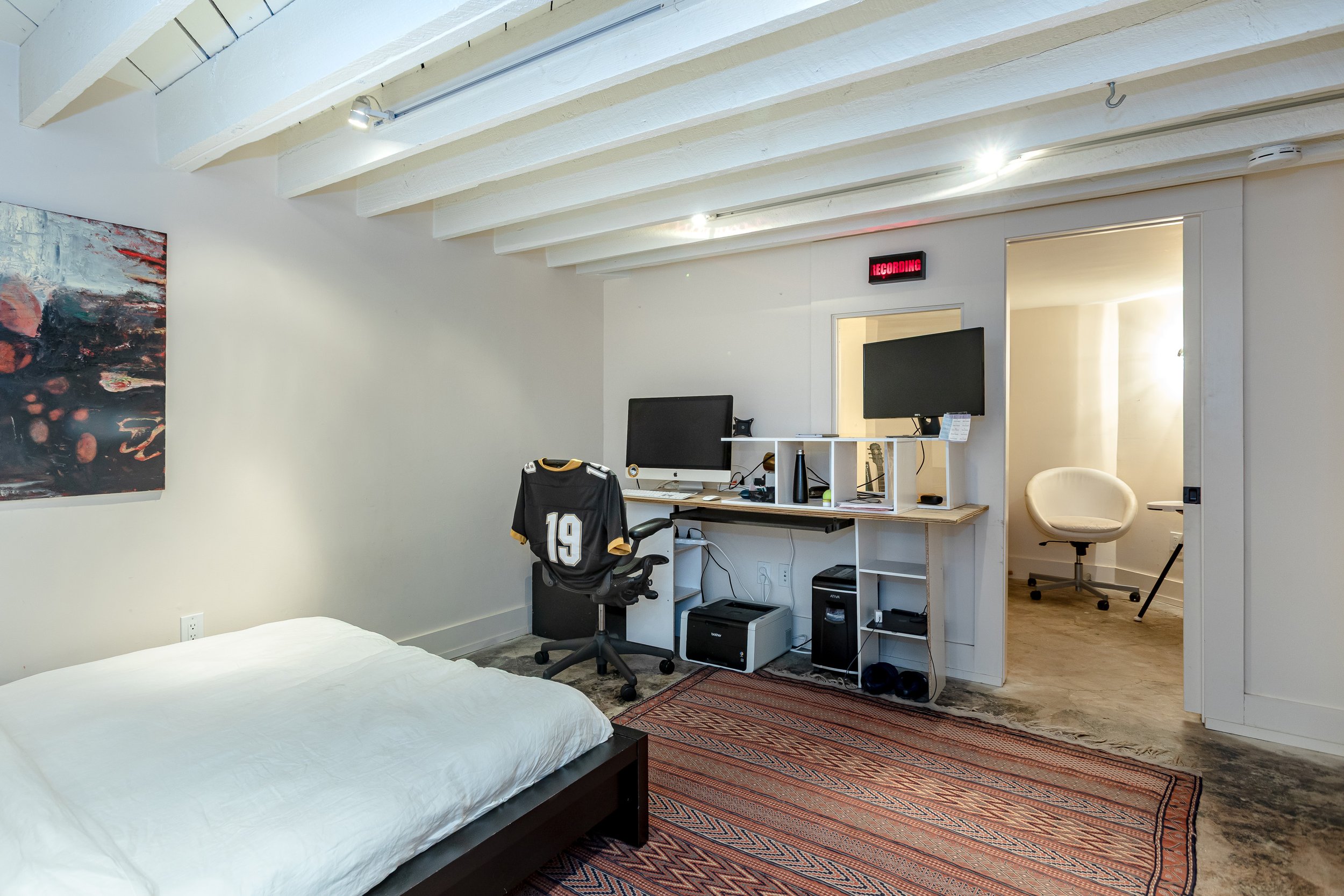 A room with a bed, a desk with two computer monitors, a black sports jersey hanging on the chair, a patterned rug, a colorful abstract painting on the wall, and a doorway leading to a smaller room with a white chair and bright sunlight.