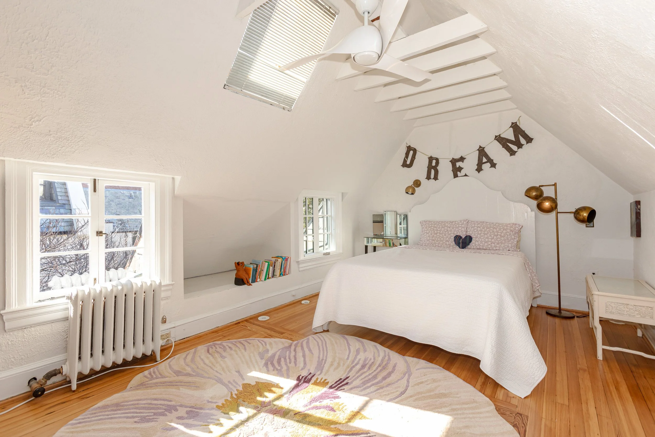 A cozy bedroom with white walls and sloped ceiling, featuring a bed with white bedding, a decorative heart pillow, and a 'DREAM' banner hanging above. There are two small windows, a white nightstand, a modern floor lamp, a round rug, and built-in she