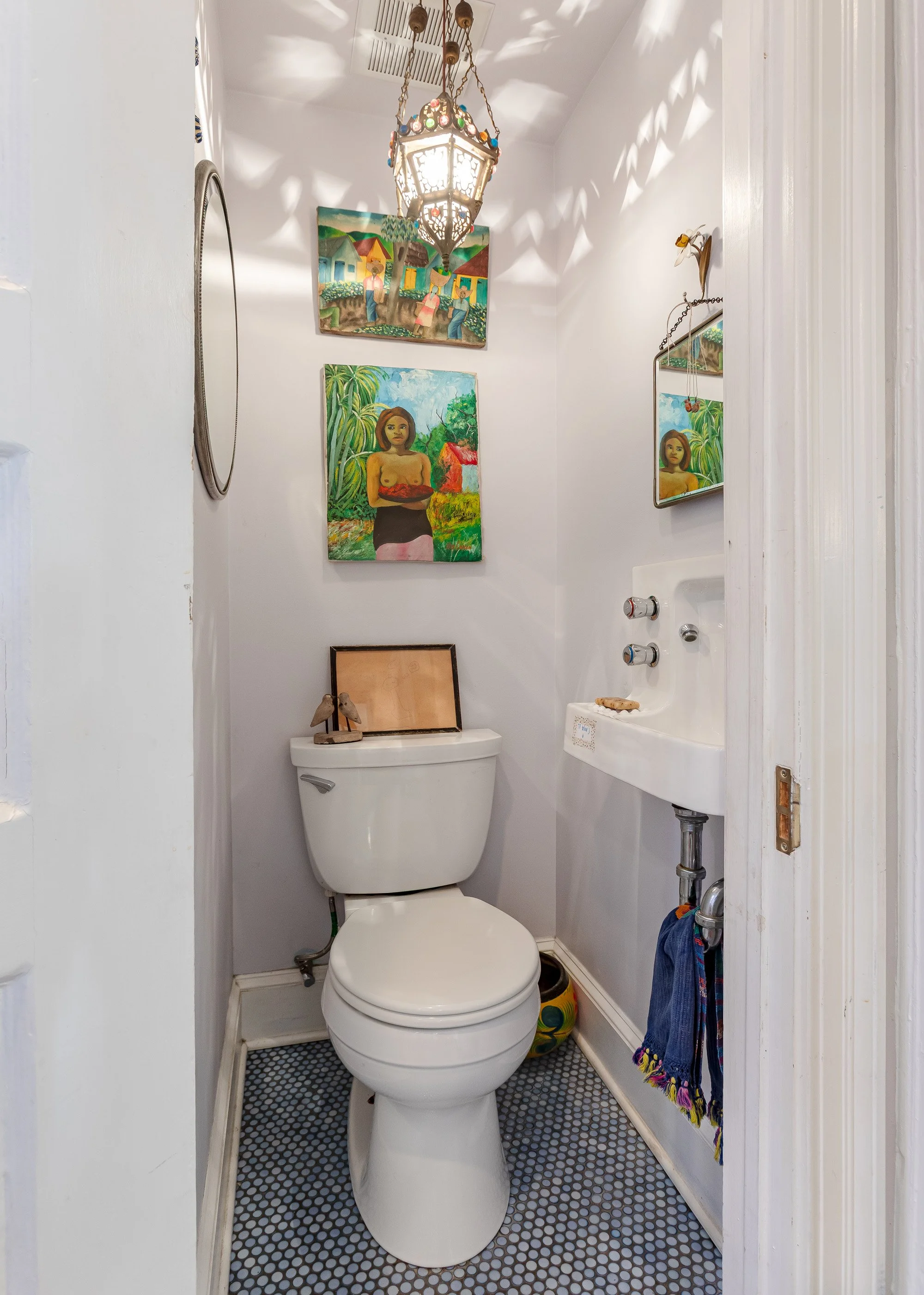 A small bathroom with a white toilet, a small wall-mounted sink, and colorful artwork on the wall. There is a decorative lamp hanging from the ceiling, casting patterned shadows, and a mirror on the right wall. The floor has small black and white til