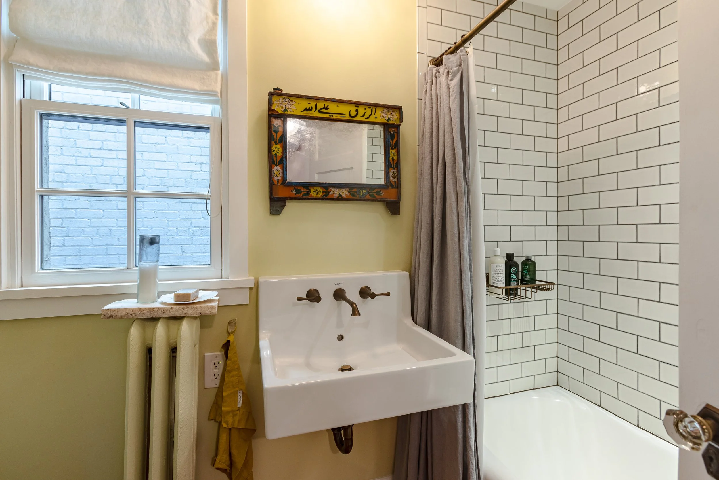 A bathroom with a window, a wall-mounted sink with a bronze faucet, a mirror with colorful floral border, a shower area with white subway tiles, shower shelf with bottles, and a curtain.