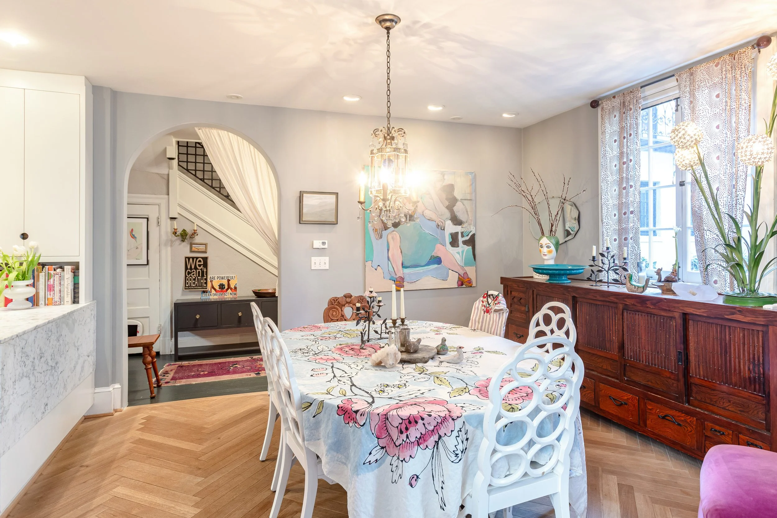 Bright dining room with a large window and patterned curtains, a wooden sideboard with decorative items, a floral tablecloth on the dining table, and a chandelier hanging from the ceiling.