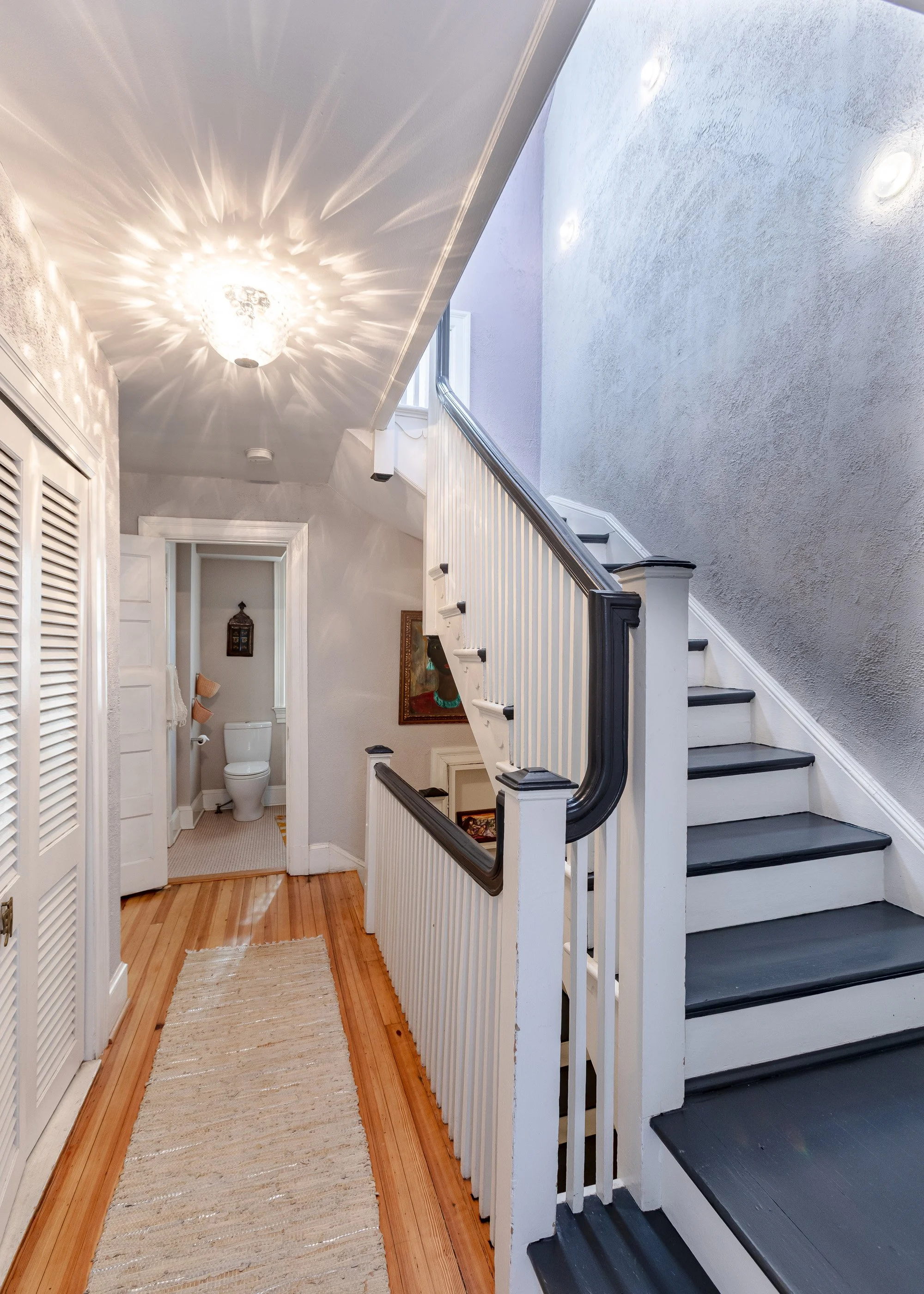 Interior view of a house hallway with a staircase, wooden floor, and a bathroom with a toilet in the background, decorated with a chandelier casting light patterns on the ceiling.
