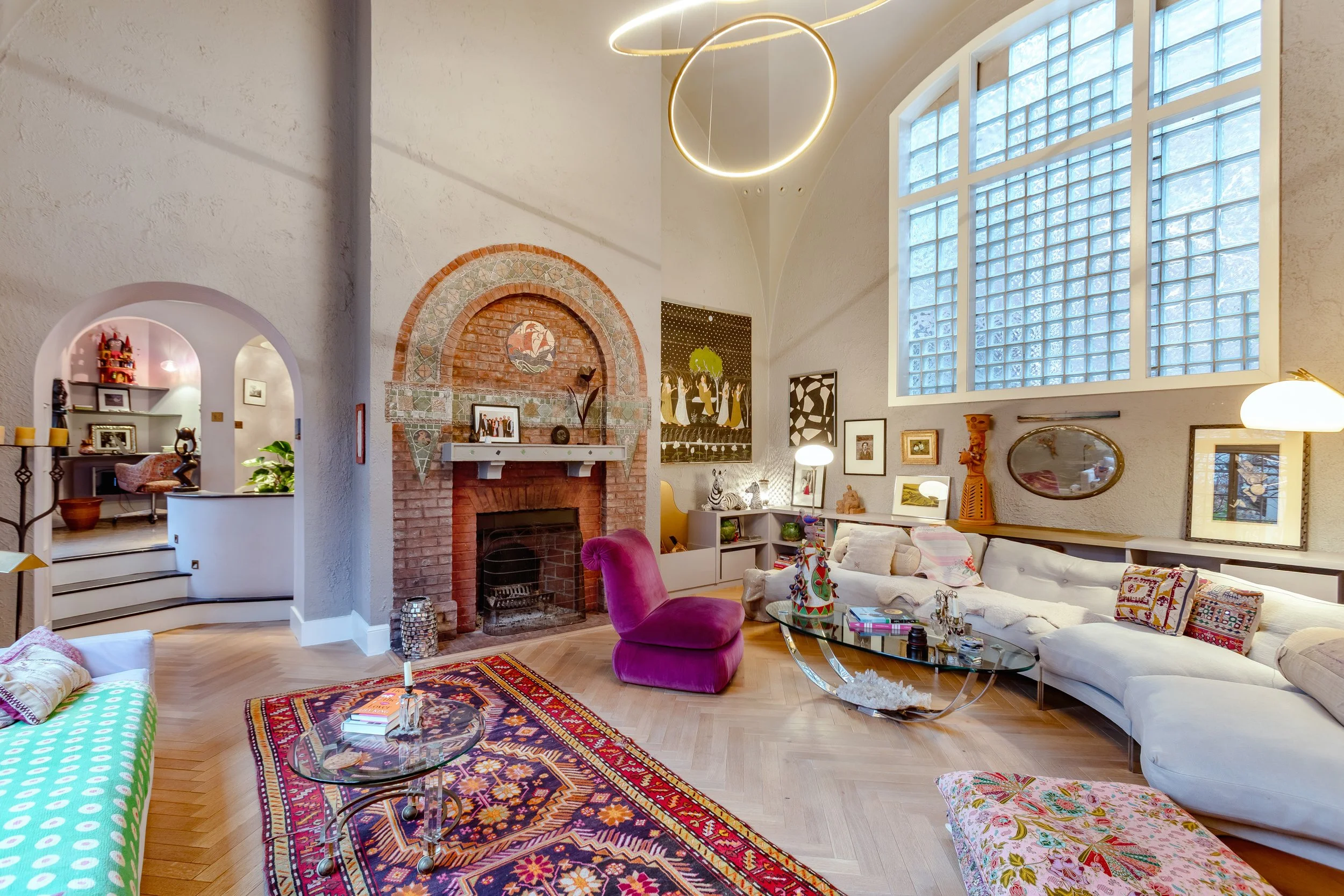 Living room with high, arched ceiling, large window, brick fireplace, colorful area rug, sectional sofa with pillows, purple velvet chair, glass coffee tables, and eclectic artwork and decorations.