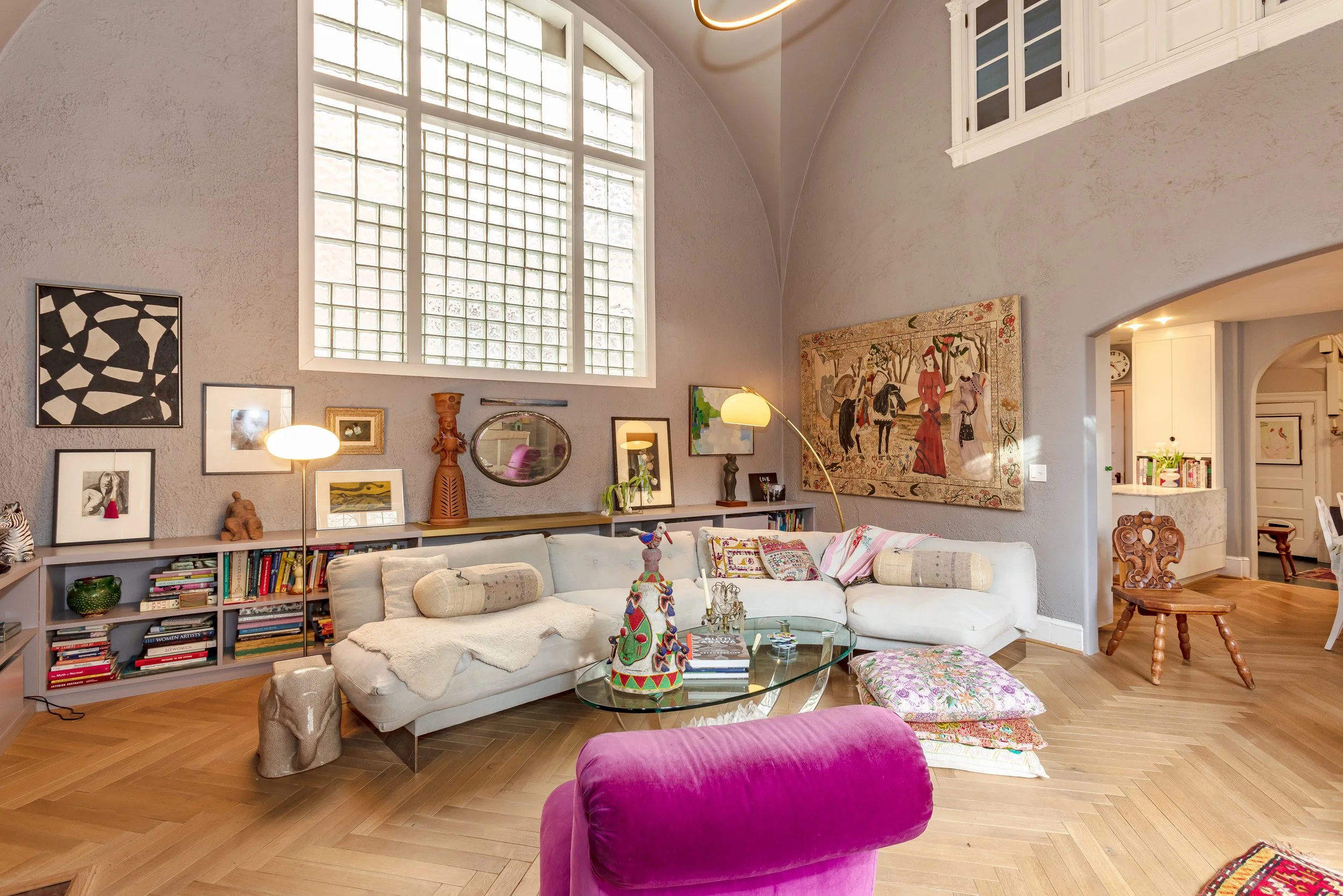 Living room with a large white sectional sofa, colorful pillows, a glass coffee table, and a pink velvet chair. The room has tall windows, artwork on the walls, a wooden chair, and hardwood floors.