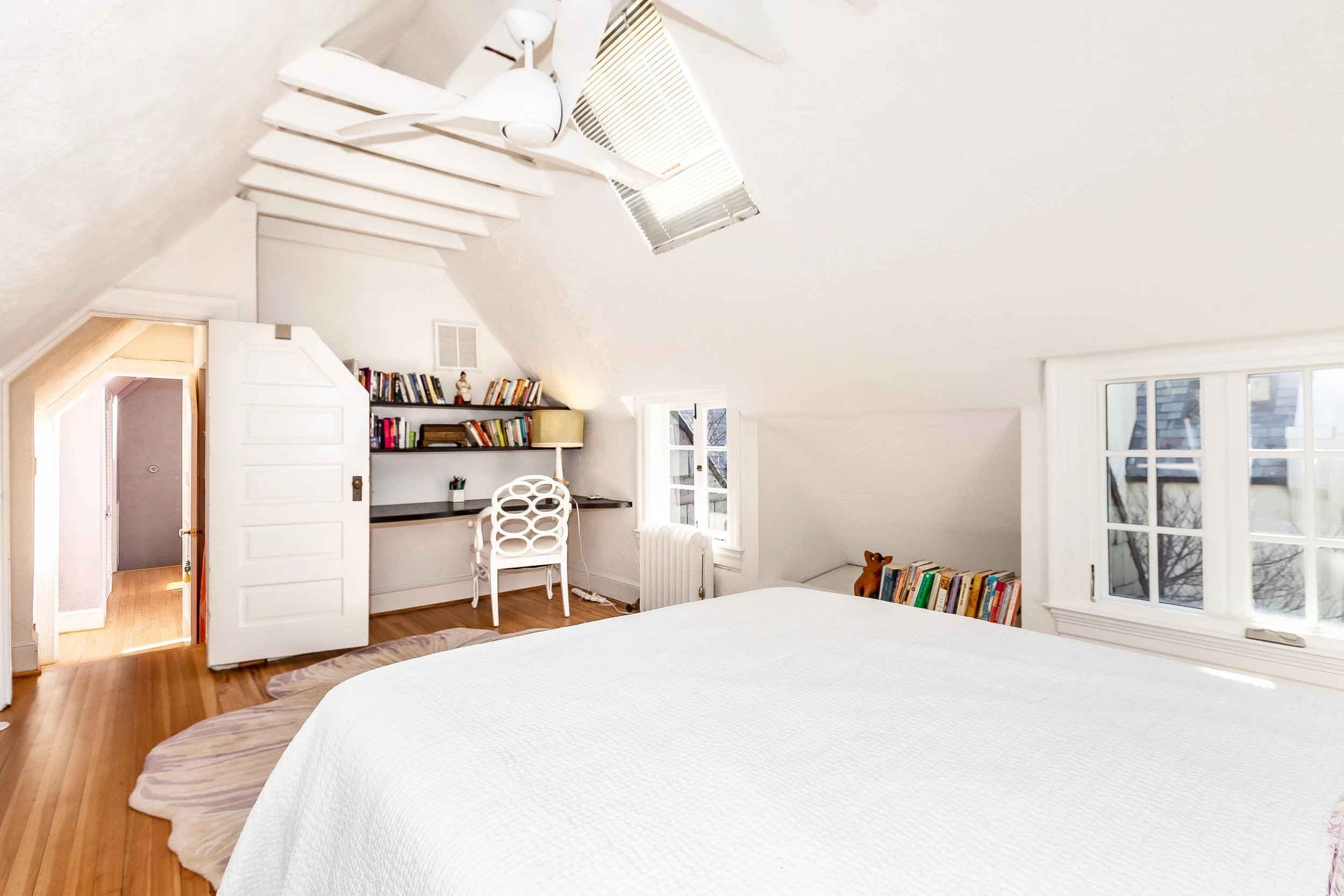 Bright bedroom with sloped ceiling, white walls, and hardwood floors. Features a bed with white bedding, a window with double panes, a small bookshelf with books, a desk with a chair, a wall-mounted shelf with books, a desk lamp, and an open door lea