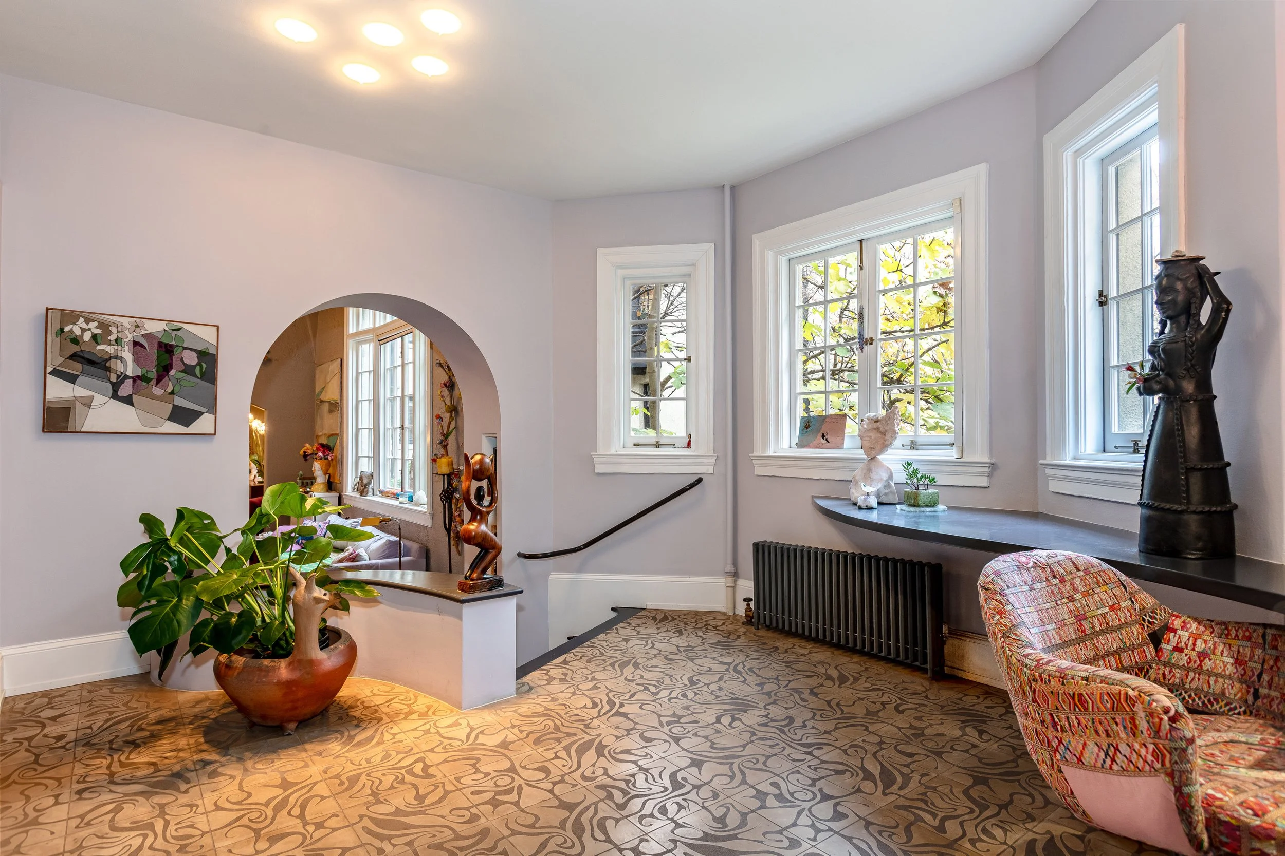 A living room with large windows, a patterned carpet, a colorful armchair, a black radiator, a shelf with sculptures and decorative items, a patterned plant in a pot, and artwork on the wall.