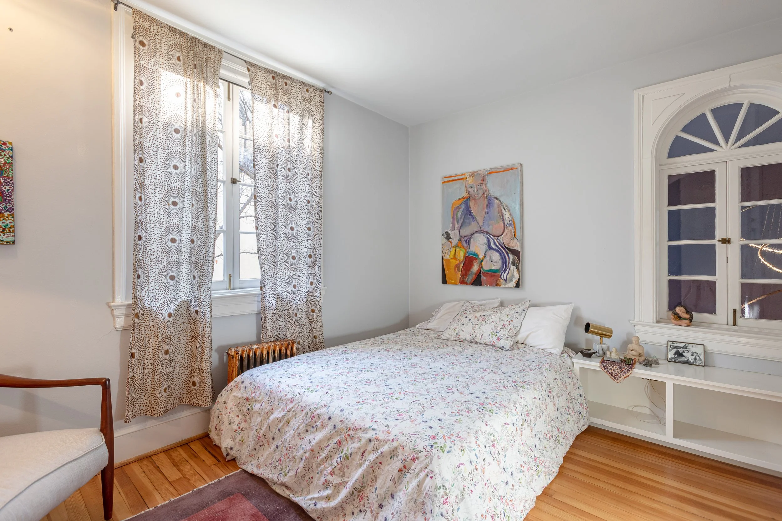 A cozy bedroom with a bed covered in a floral duvet and pillows, artwork on the wall, a side table with decorative items, a window with patterned curtains, and a chair next to the bed.