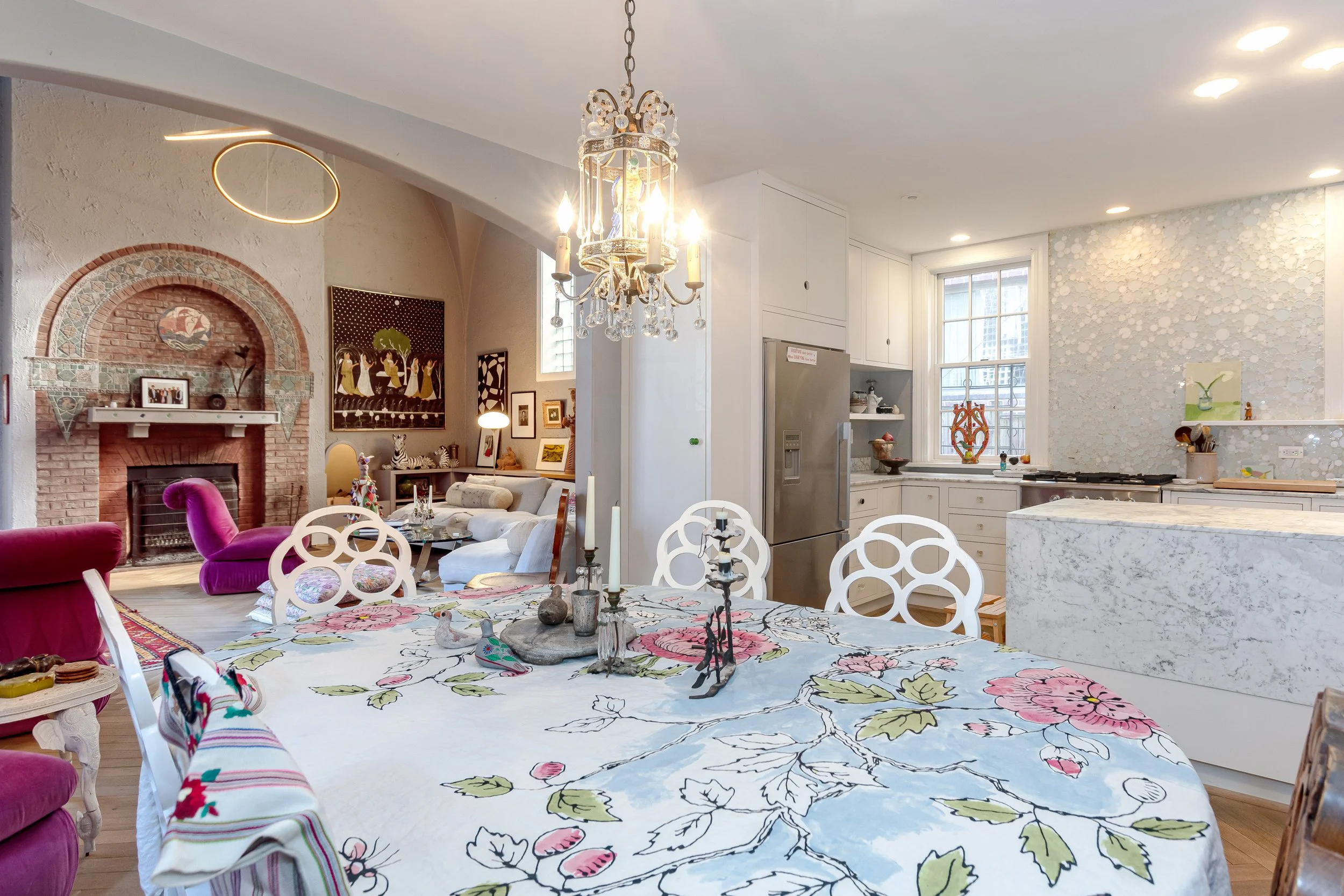 Open-concept kitchen and dining area with a chandelier, floral tablecloth, and colorful chairs. Living room in the background with a purple armchair, white couch, and artwork above a brick fireplace.