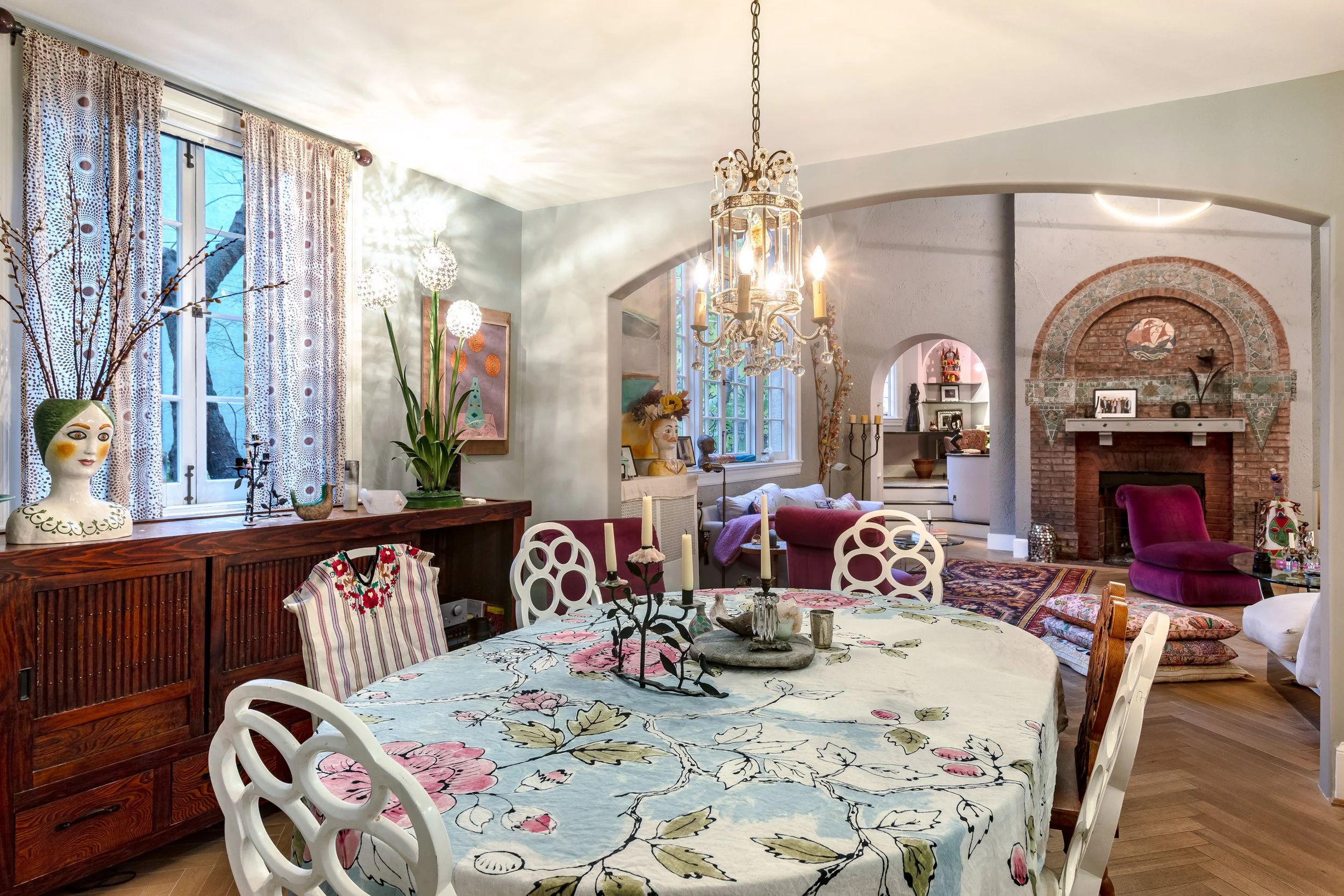 A decorated living and dining room with a wooden sideboard, floral tablecloth, chandelier, colorful artwork, large window with patterned curtains, and a brick fireplace surrounded by decorative pillows and vintage furniture.